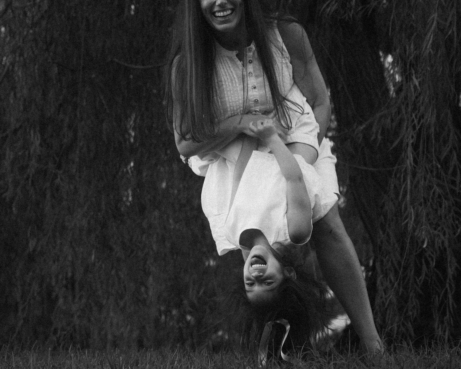Black and white photo of two girls laughing and playing outdoors, one held upside down joyfully.