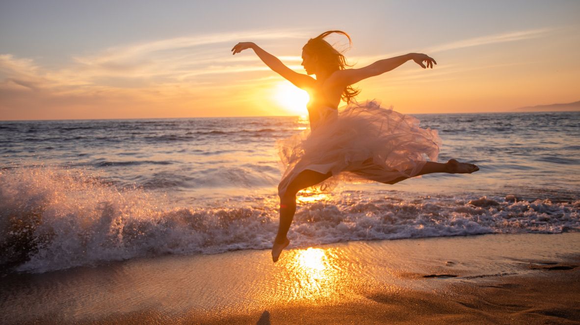 A silhouetted figure leaps through ocean spray at sunset on the beach.