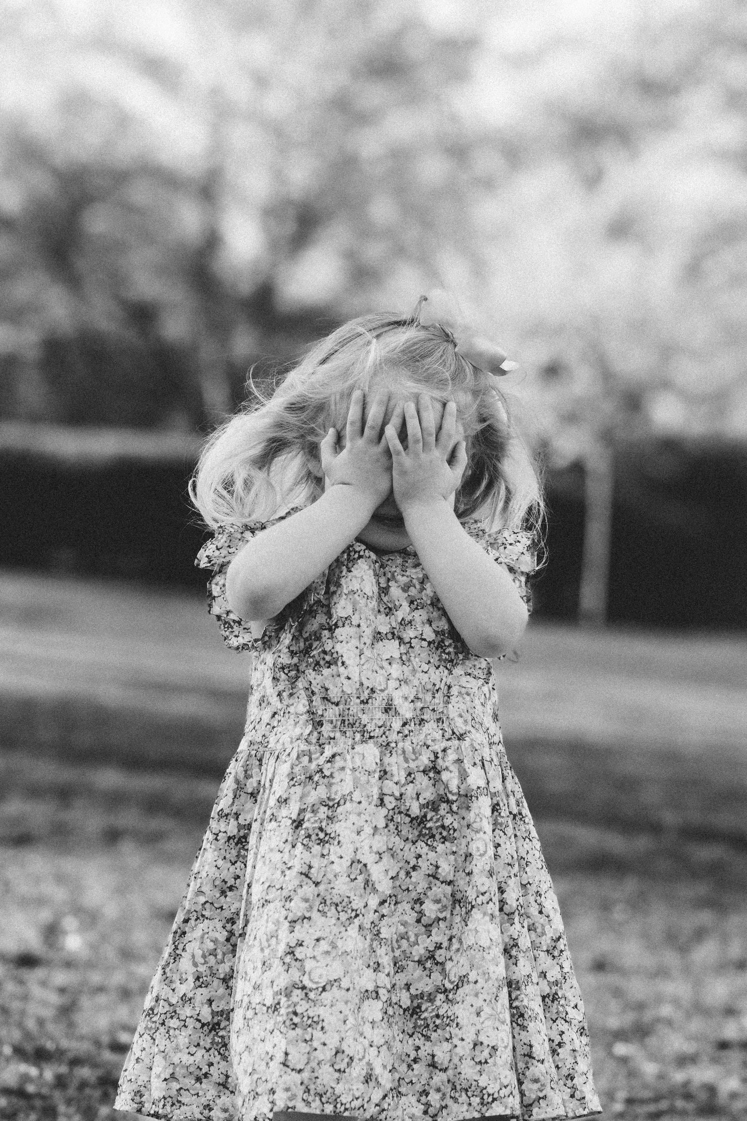 Black and white photo of a young girl in a floral dress covering her face with both hands outdoors.