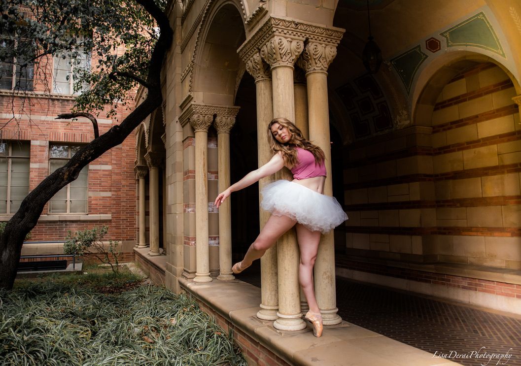 Dancer in white tutu performs graceful movements under ornate stone archways in dramatic lighting.