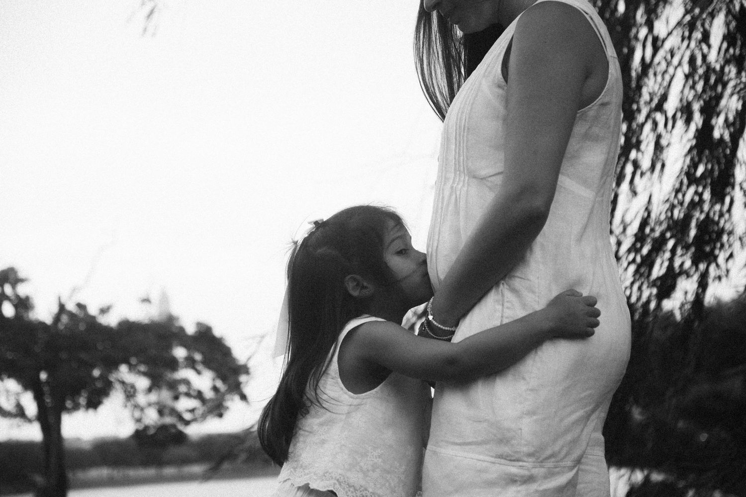 Black and white photo of a young girl hugging her pregnant mother outdoors near trees.