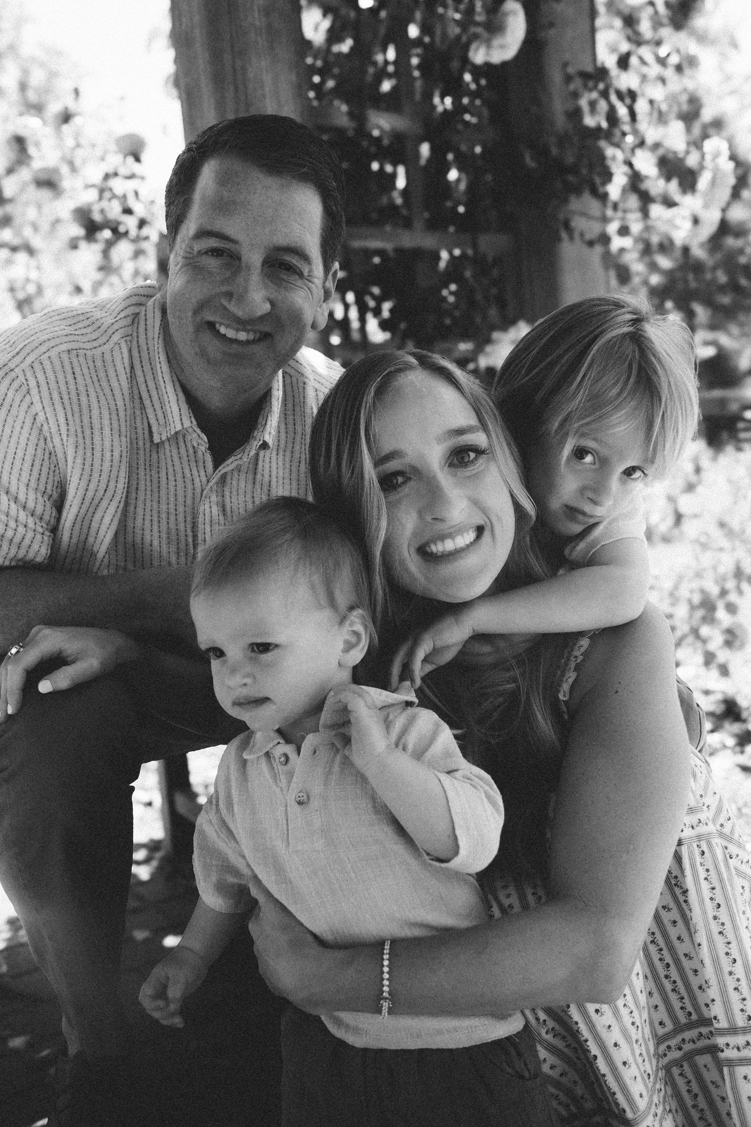 Black and white family portrait of parents smiling with two young children outdoors under a tree.