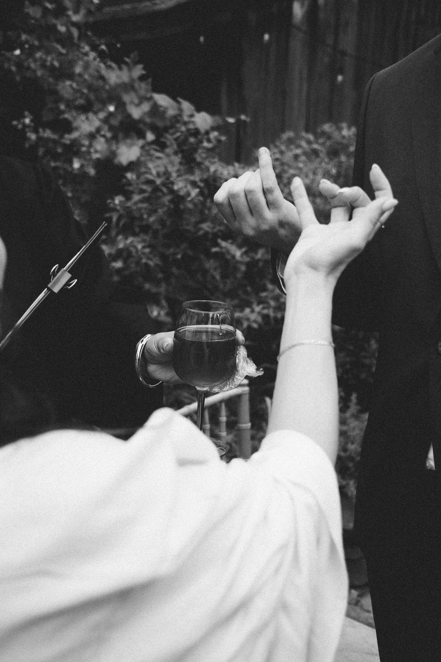 Black and white photo of a bride holding wine glass reaching up to touch groom's hand at outdoor wedding ceremony.