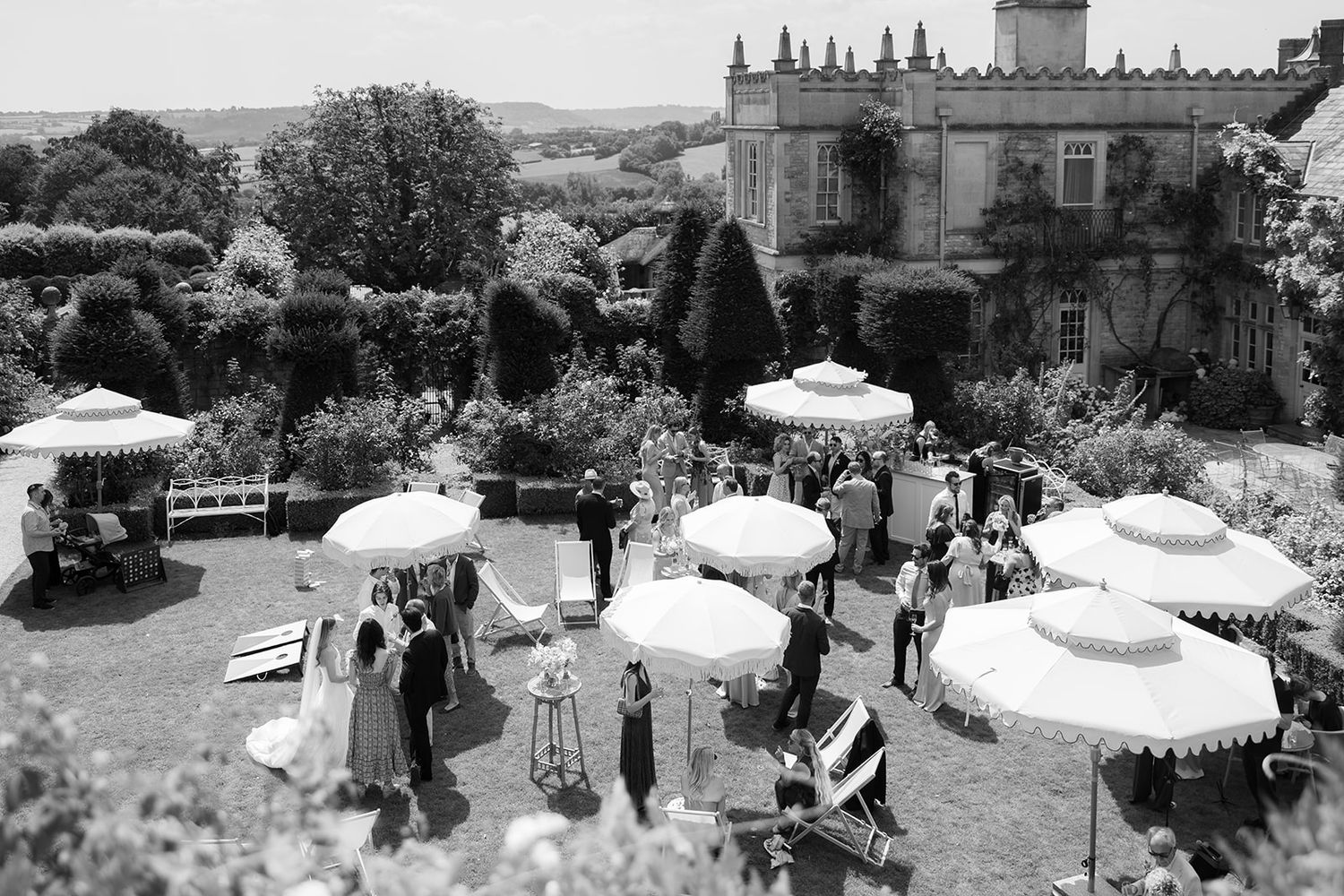 Black and white aerial view of an elegant garden party with white parasols on a manicured lawn beside a historic manor.