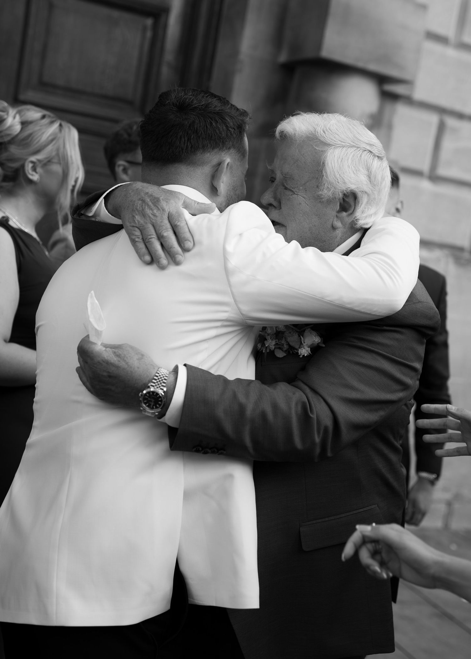 Black and white photo of two men sharing an emotional hug at a wedding ceremony outdoors.