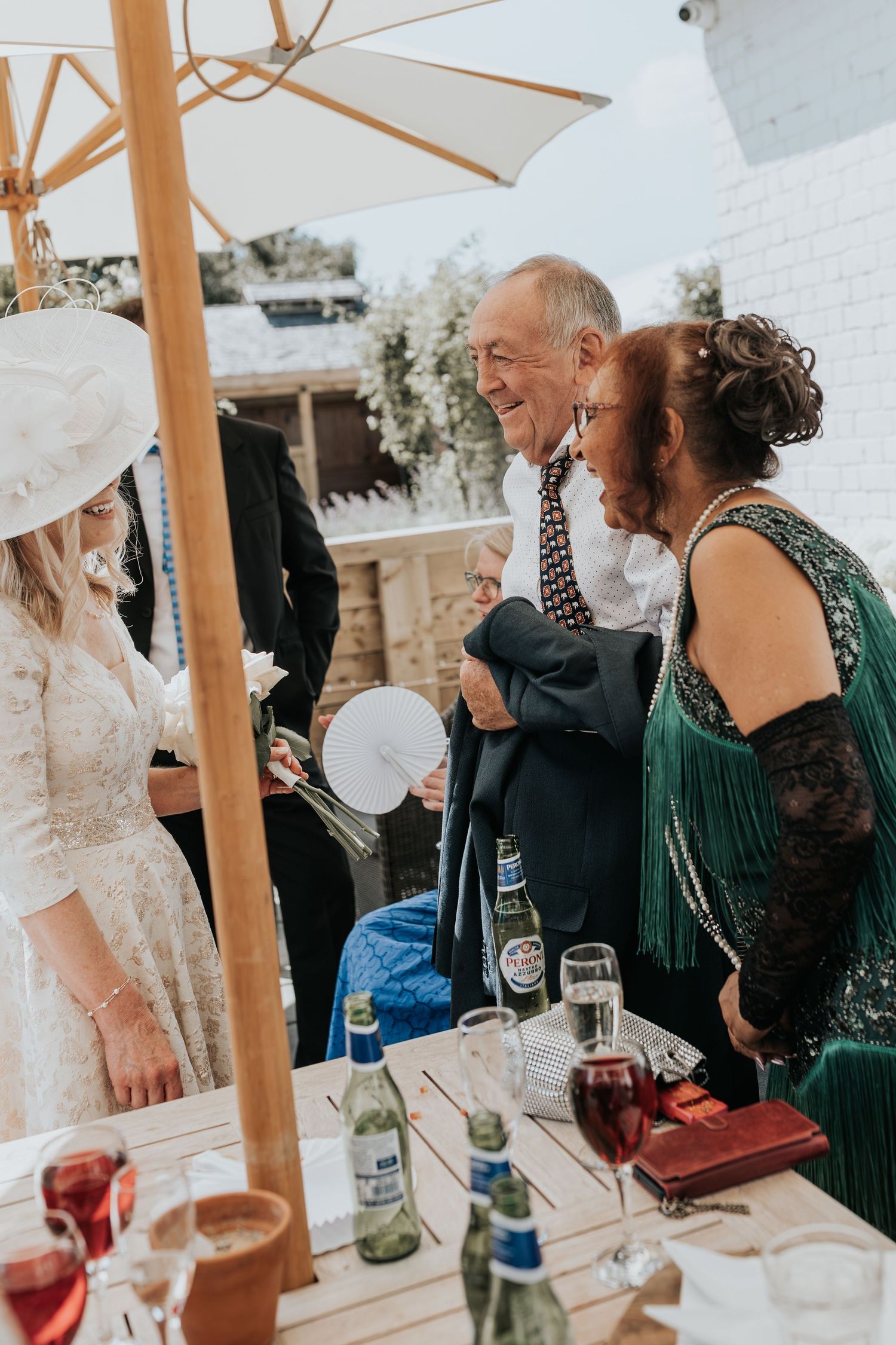 Wedding guests in elegant attire socializing outdoors, woman in white lace dress and hat, man in suit, woman in green dress.