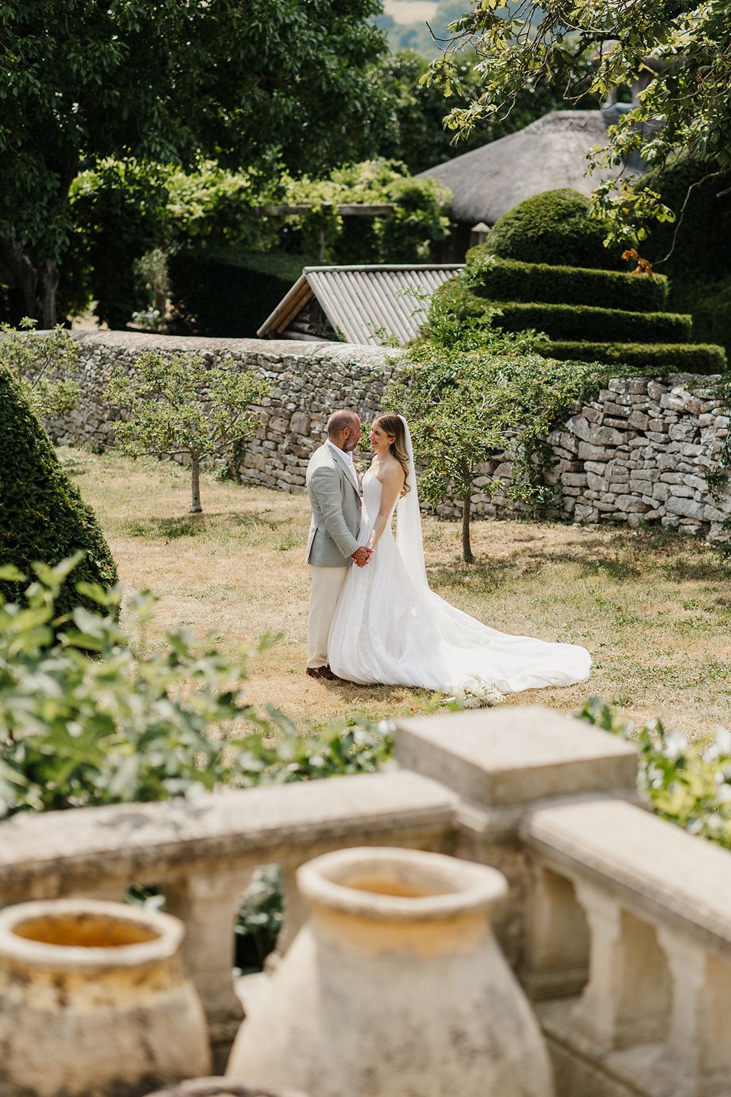 A bride and groom share an intimate moment in a sunlit French garden with stone walls and manicured hedges.
