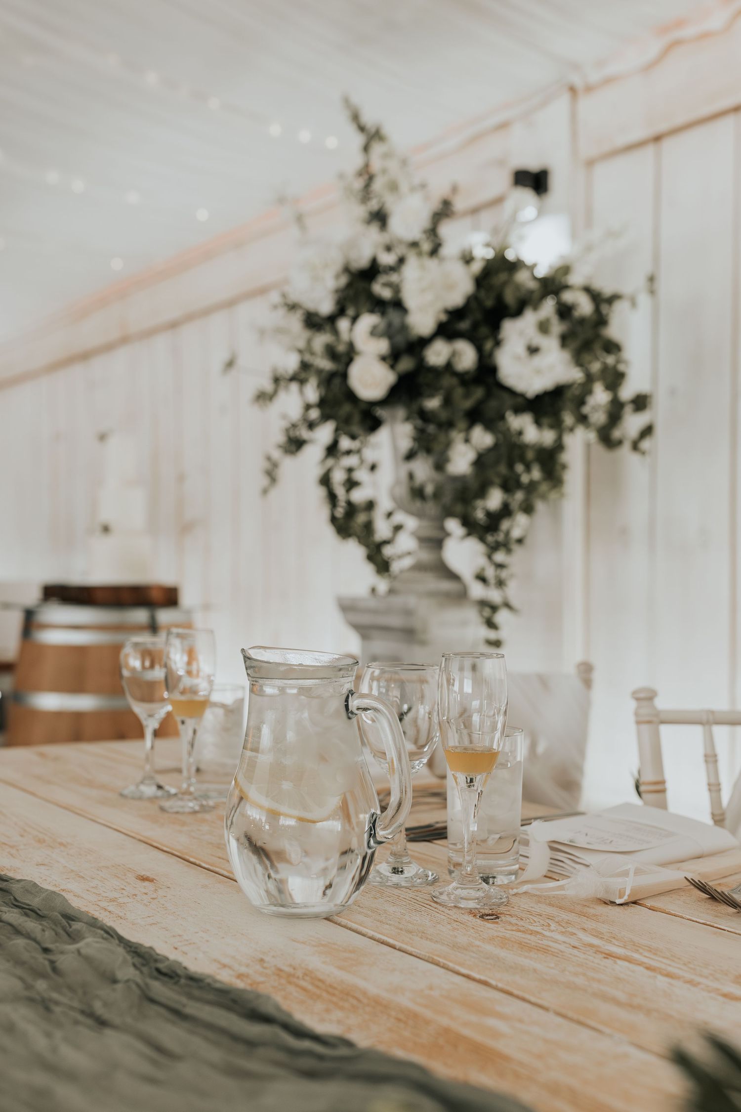Elegant wedding reception table with glass pitcher, stemware, and lush white floral centerpiece in a white tent venue.