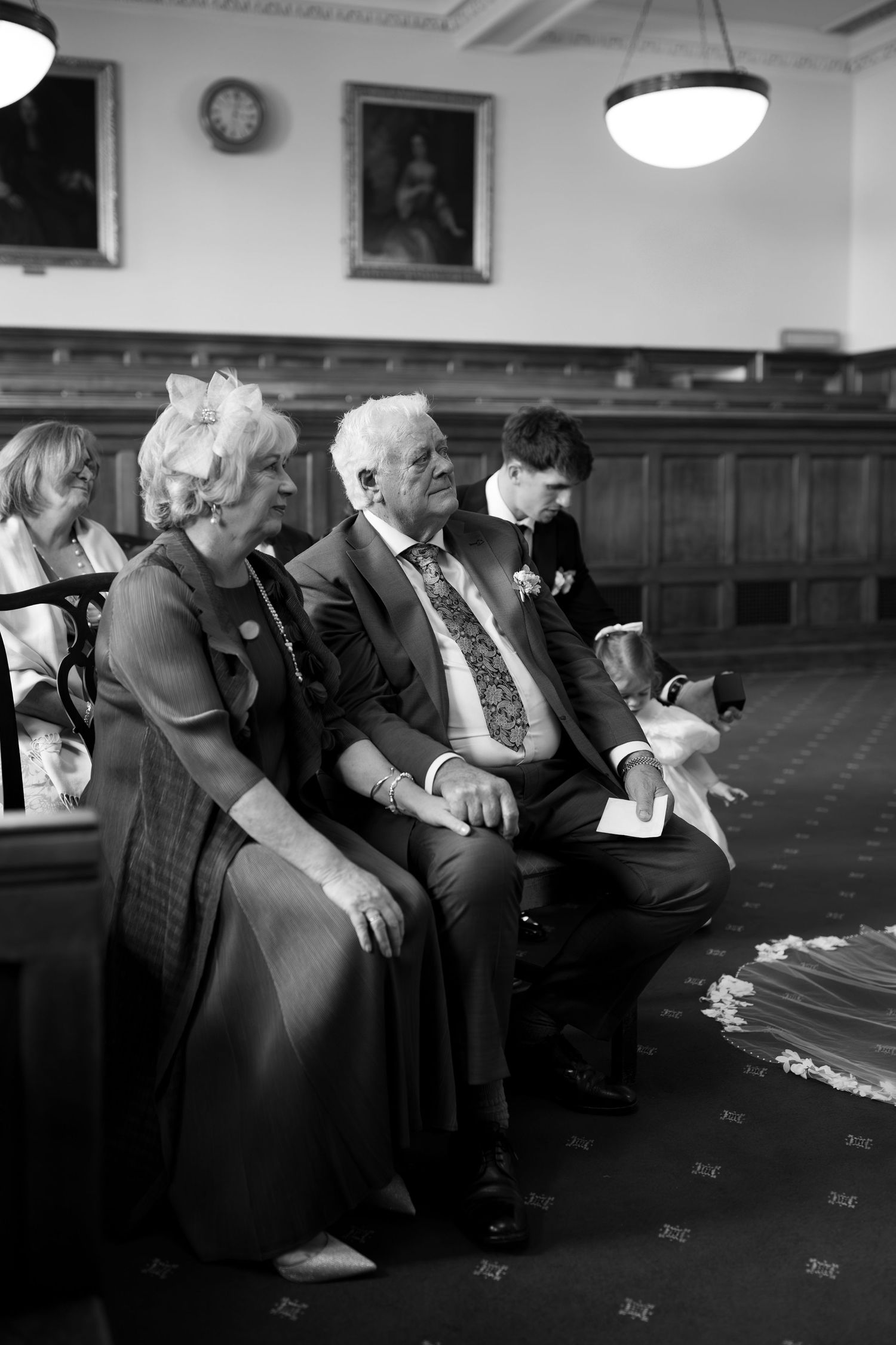 Black and white photo of elderly wedding guests seated in a formal hall, watching a ceremony with a chandelier above.