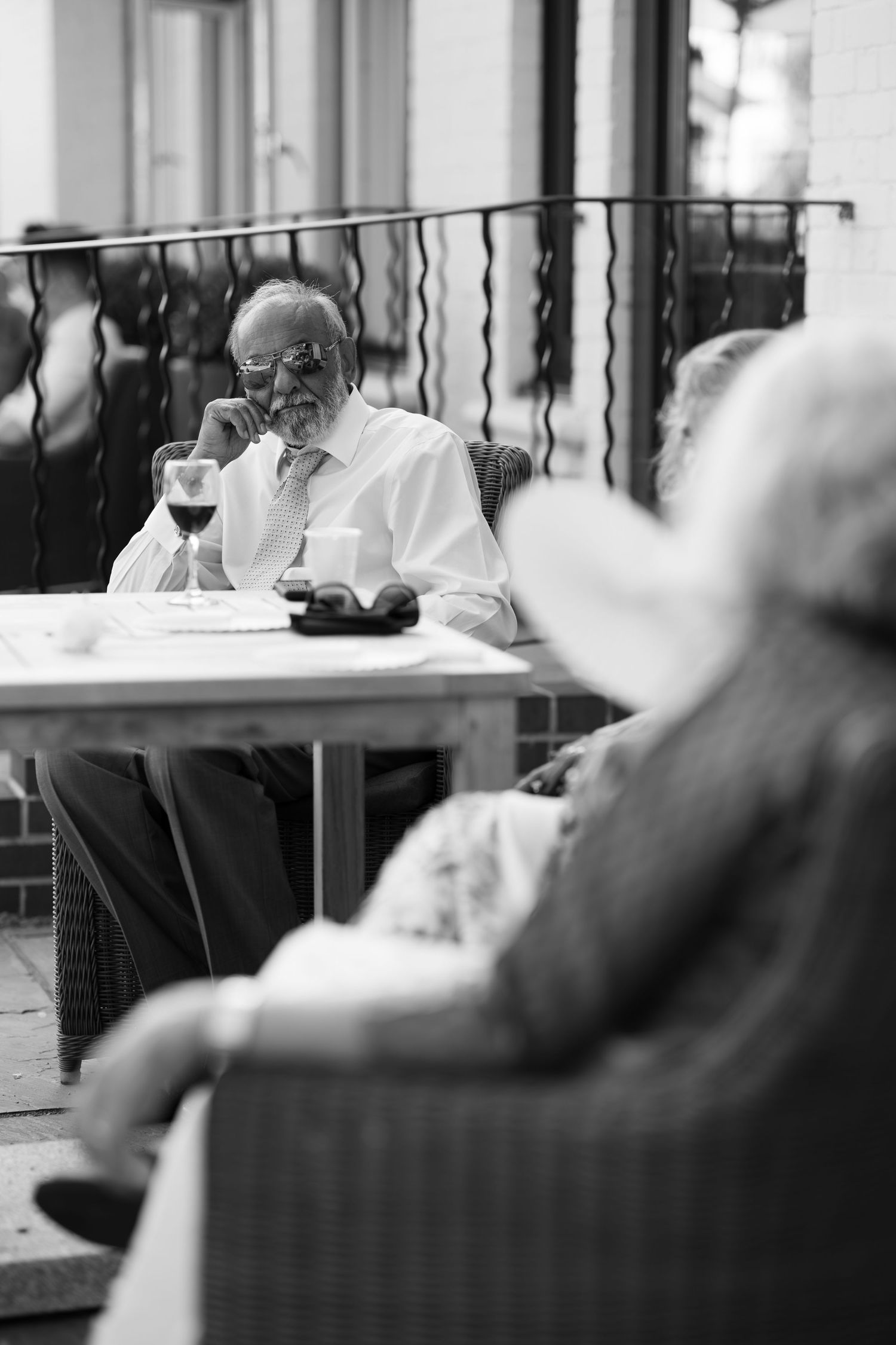 Black and white photo of an elderly man in glasses enjoying a glass of wine at an outdoor cafe table.