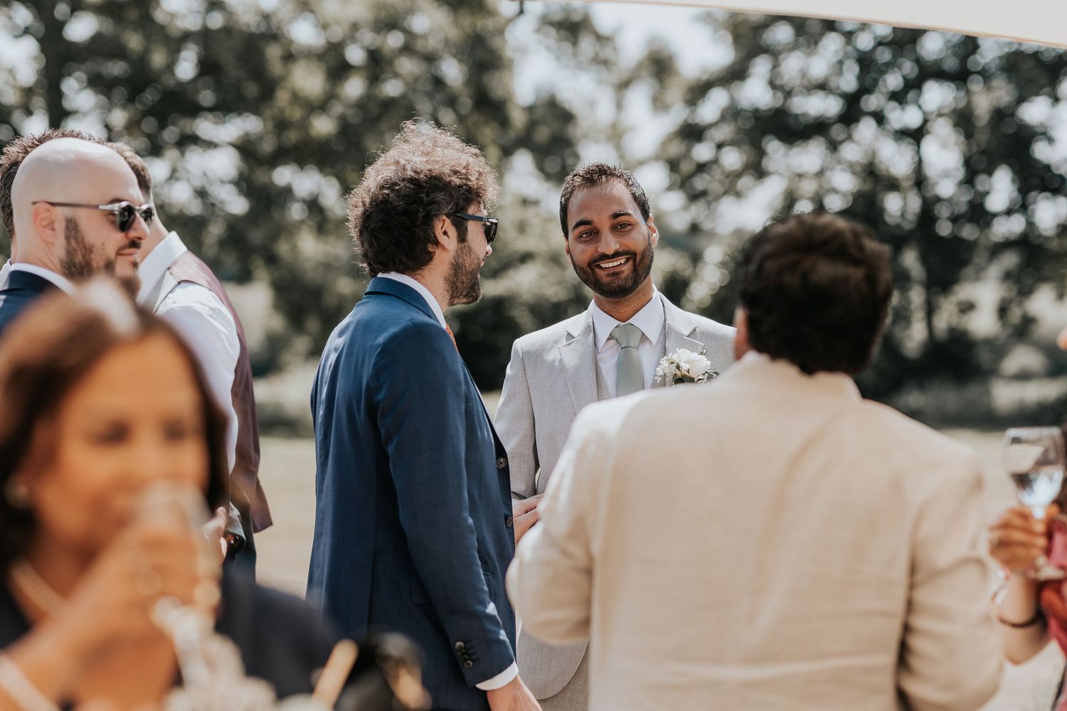 Groom in light suit smiling at wedding guests outdoors, surrounded by men in suits at a garden celebration.