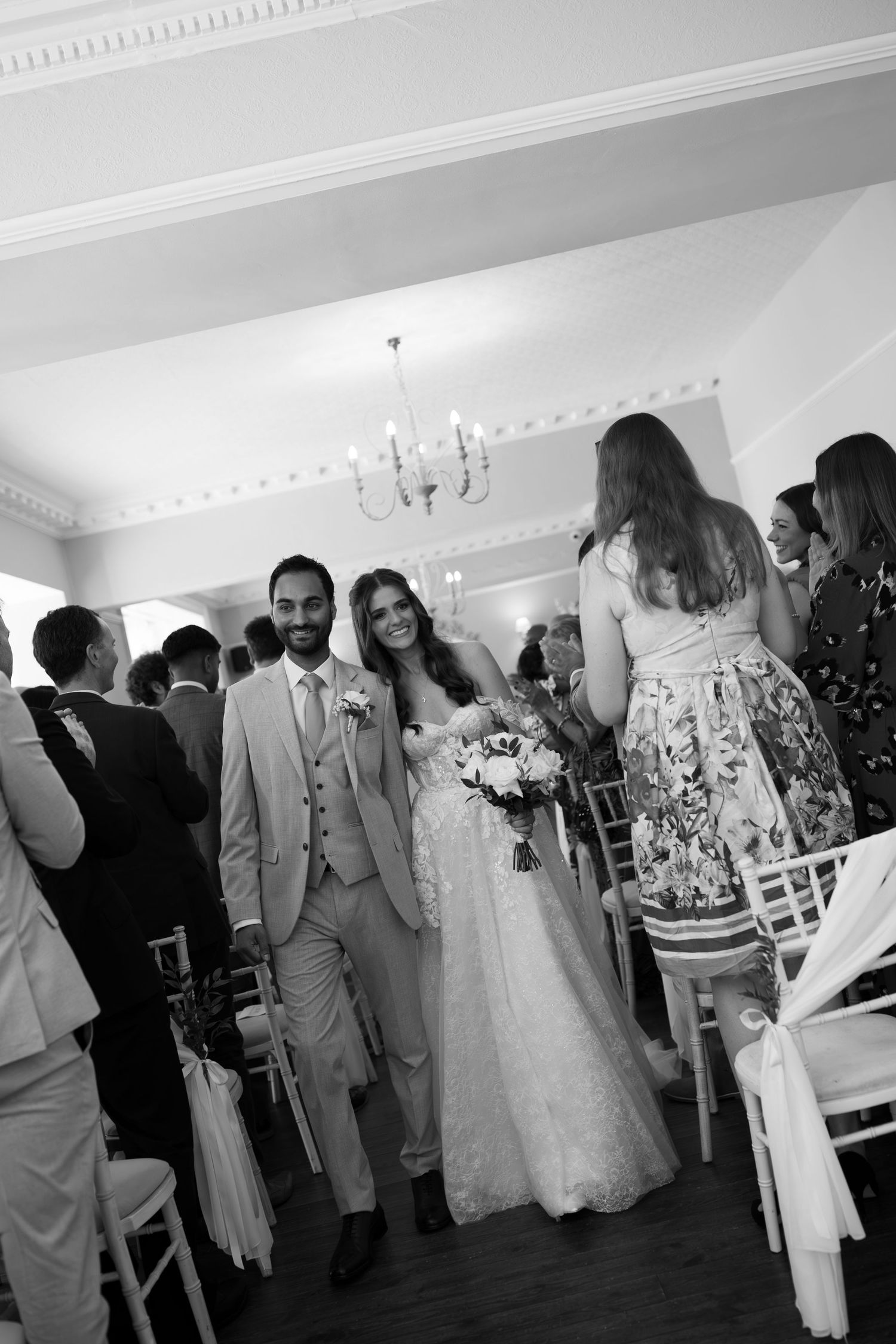 Black and white photo of a smiling bride and groom walking down the aisle at their elegant indoor wedding ceremony.