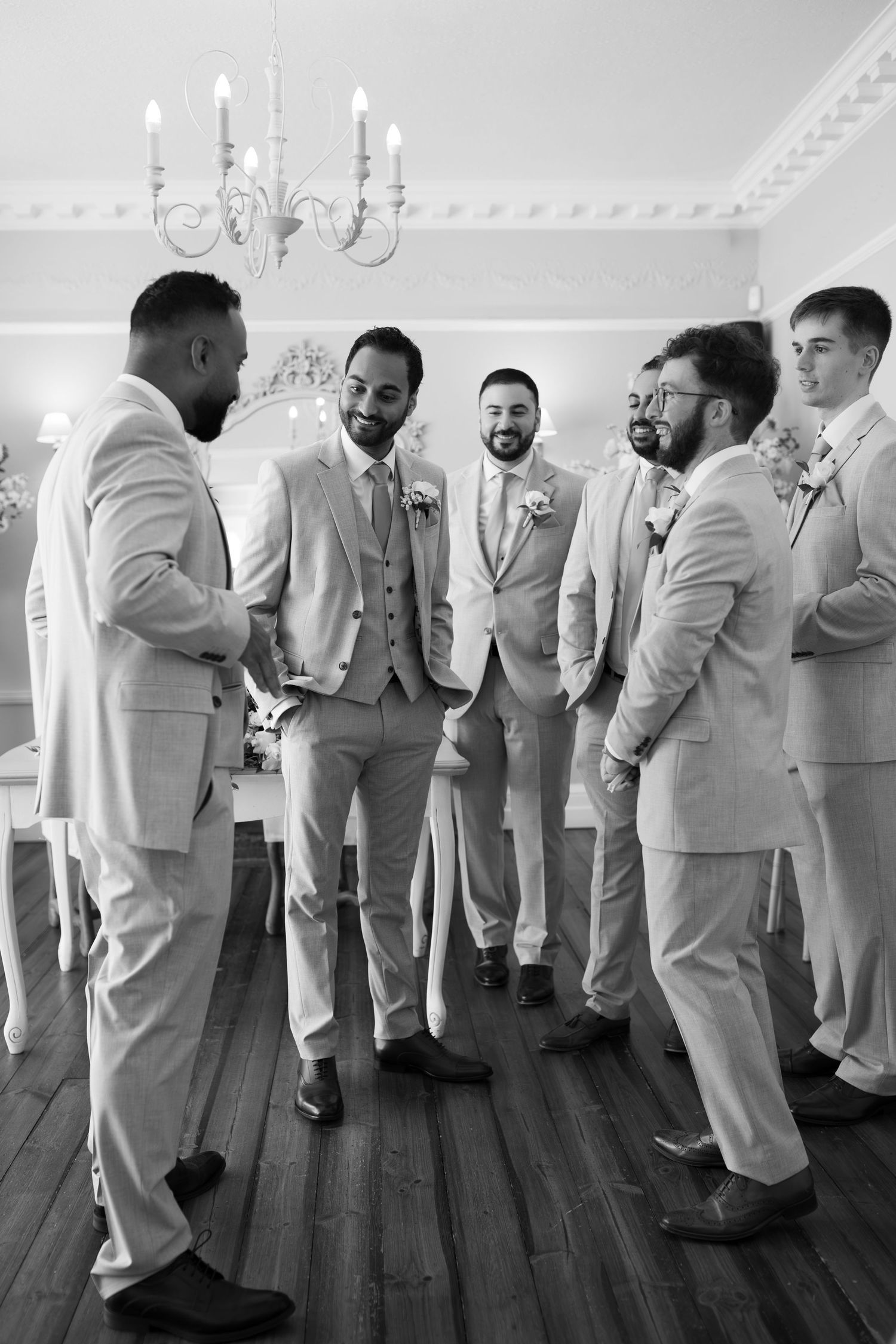 Groom and groomsmen in light suits laughing together in elegant room with chandelier, black and white wedding photo.