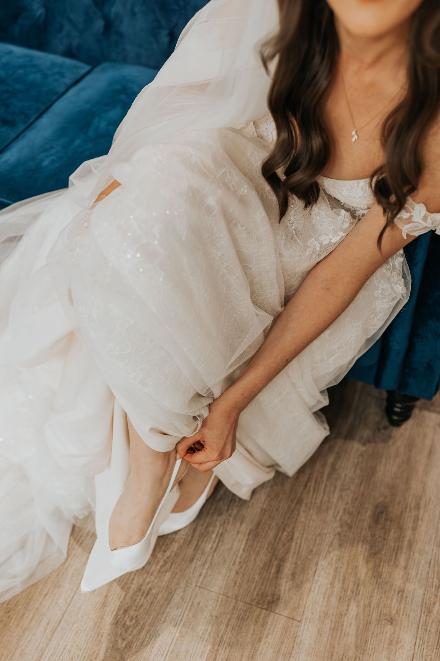 Bride in white tulle wedding gown sitting on blue velvet sofa, putting on white satin heels on hardwood floor.