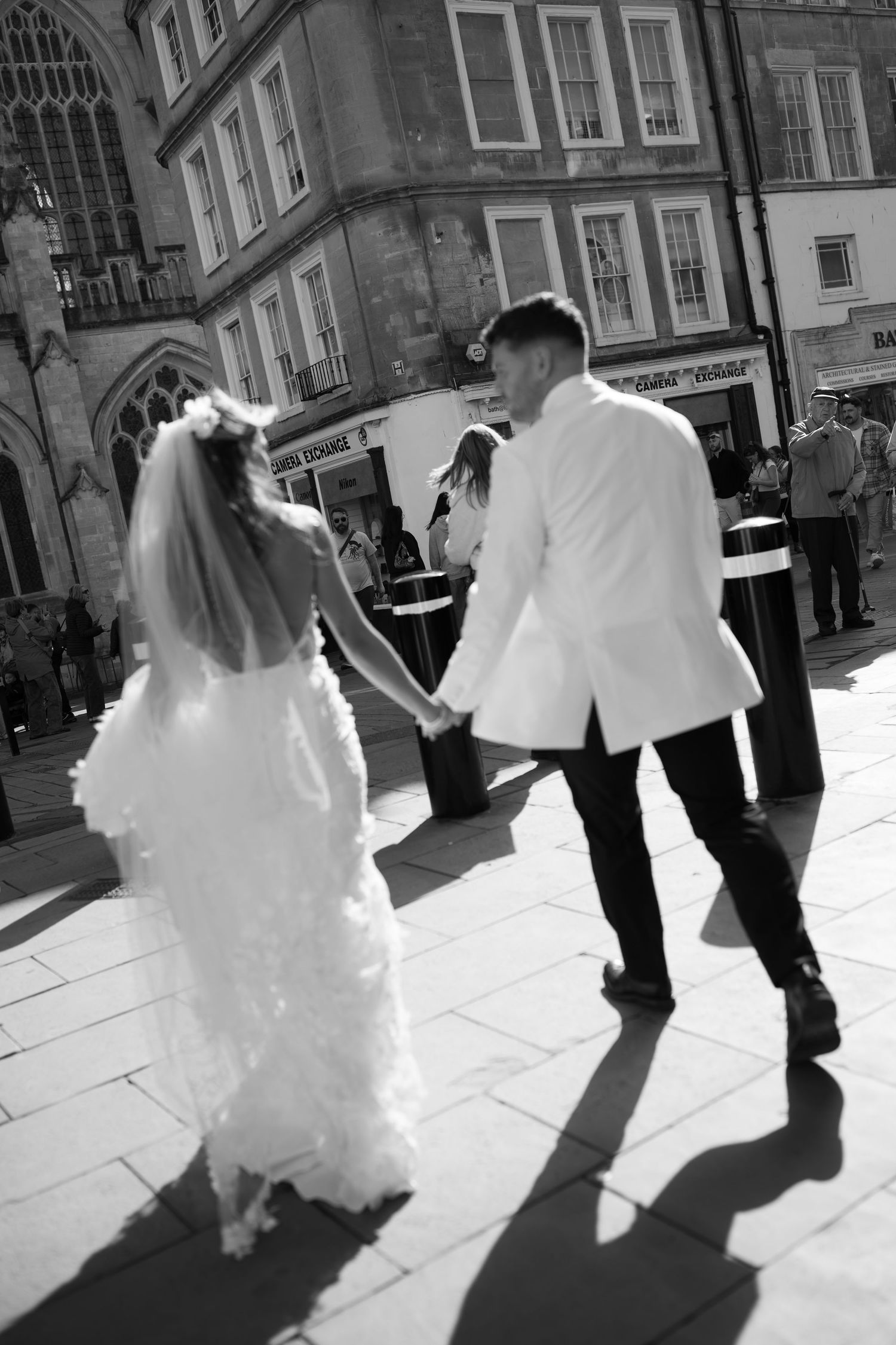 Black and white photo of a bride and groom holding hands, walking through a sunny cobblestone plaza on their wedding day.