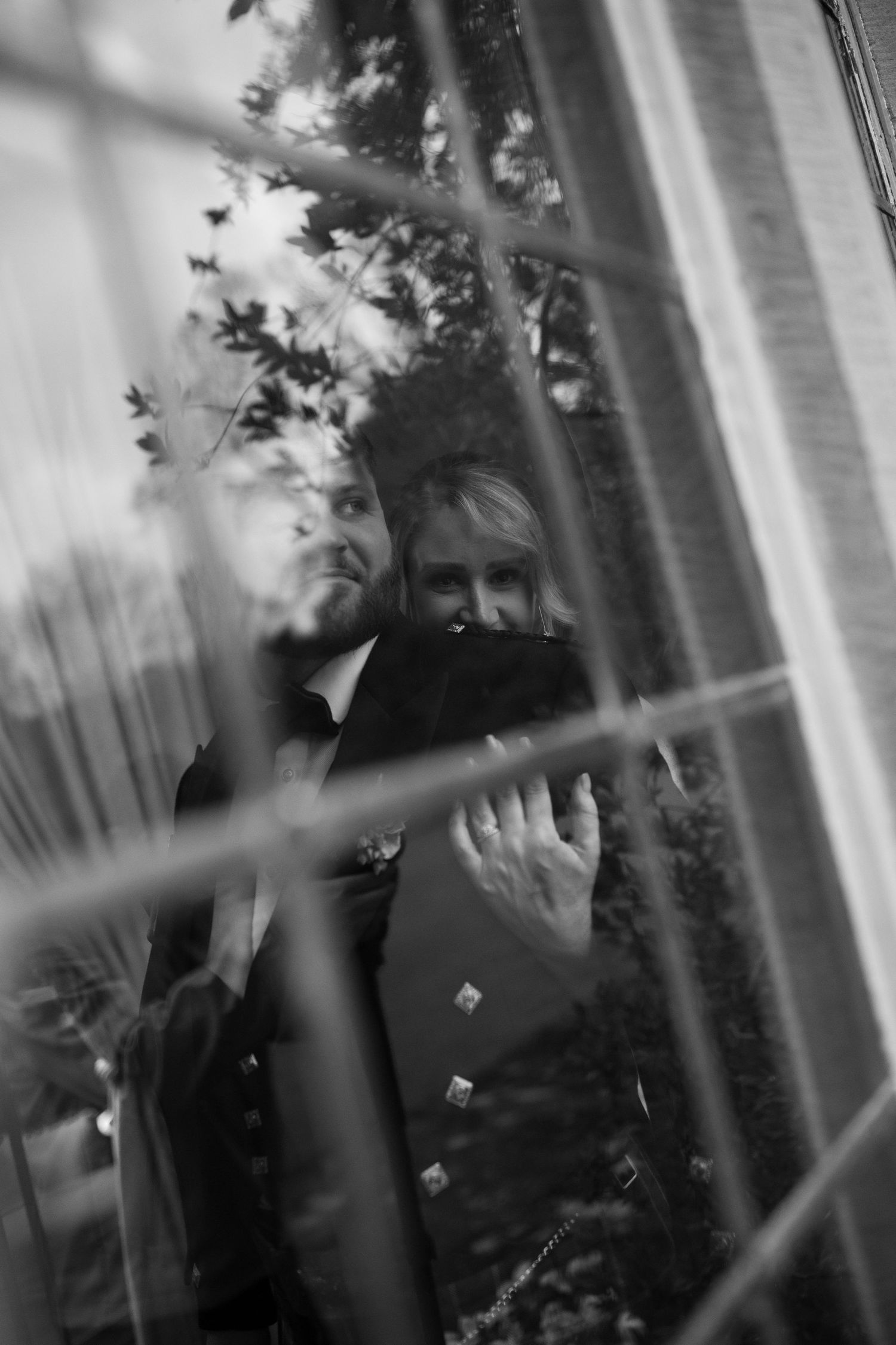 A black and white photograph of a person's face partially obscured by a window or glass surface, with foliage visible in the background.