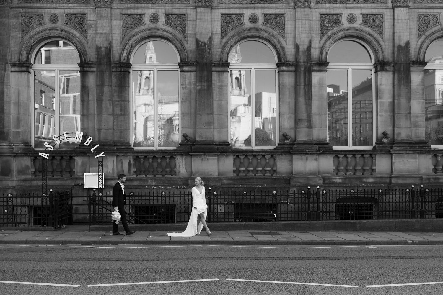 Black and white photo of a bride and groom walking past a grand stone building on a city street.