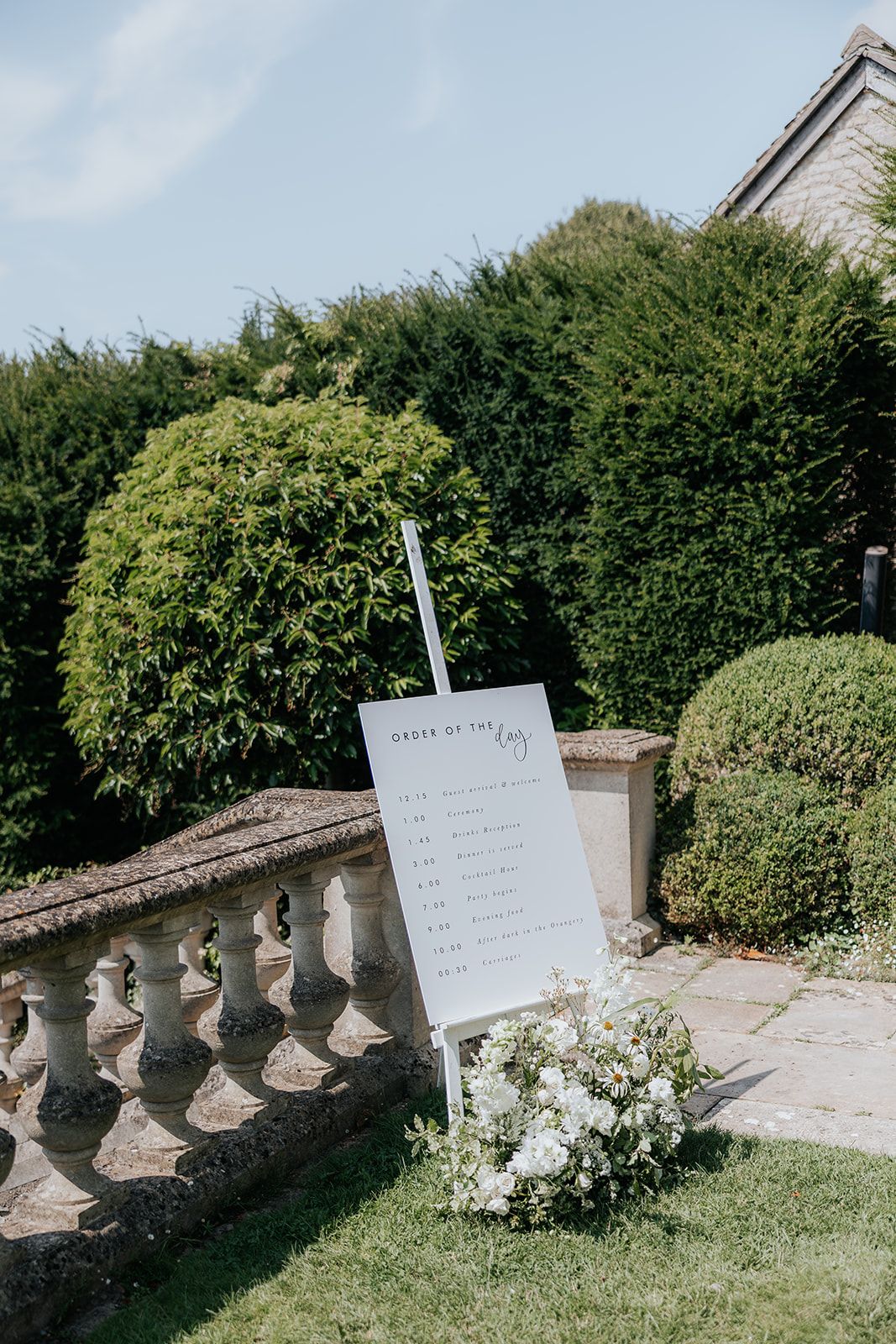 Elegant outdoor wedding order of the day sign on a white easel with floral arrangement, set against lush green hedges.