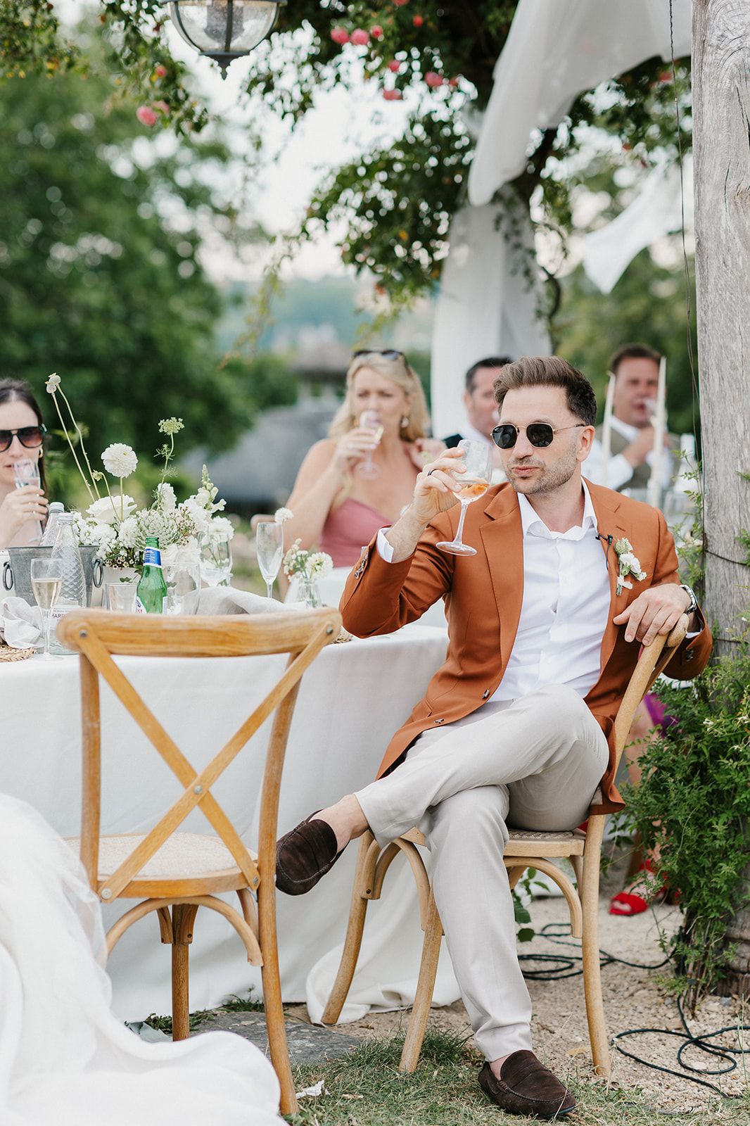 Stylish groom in rust blazer and sunglasses holds a cocktail at an elegant outdoor wedding reception.