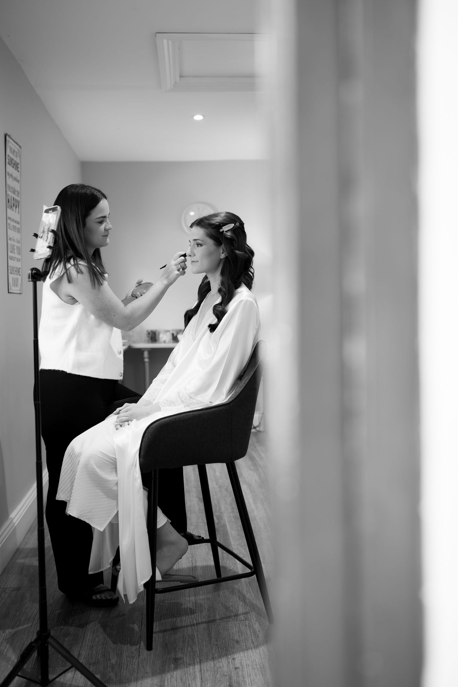 Black and white photo of a bride getting makeup applied by a makeup artist in a bright room before her wedding.