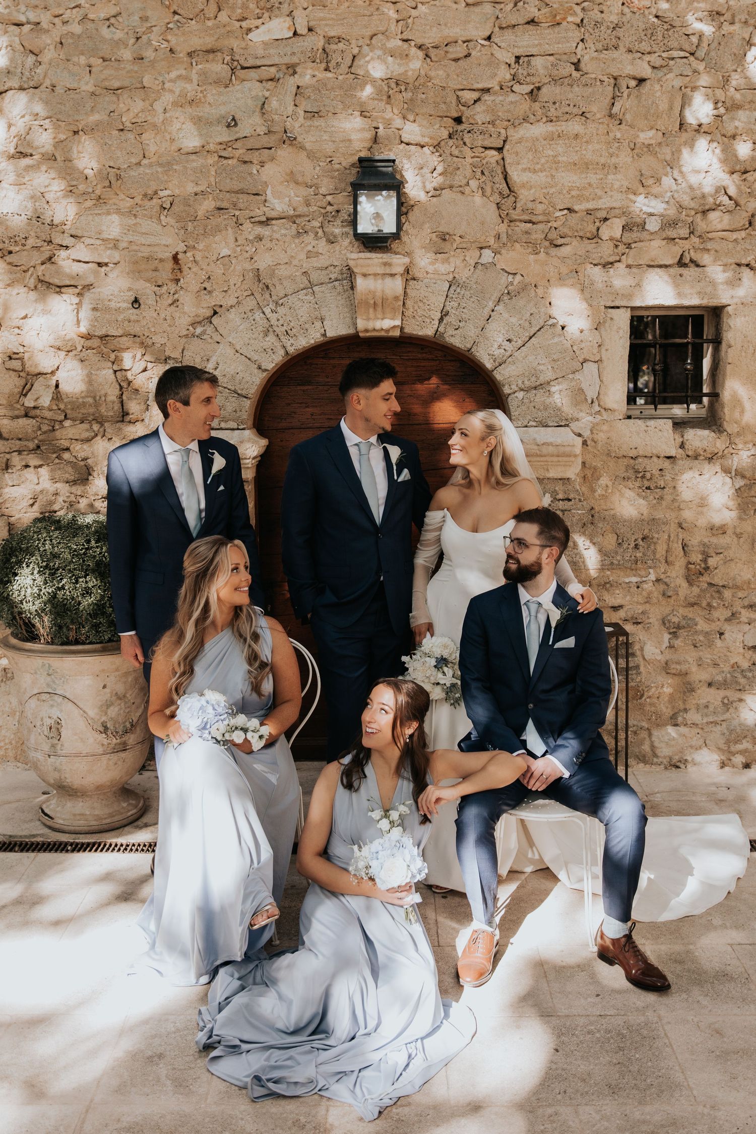The image shows a group of people dressed in formal attire posing for a wedding photo in front of a stone wall with an archway and lantern.