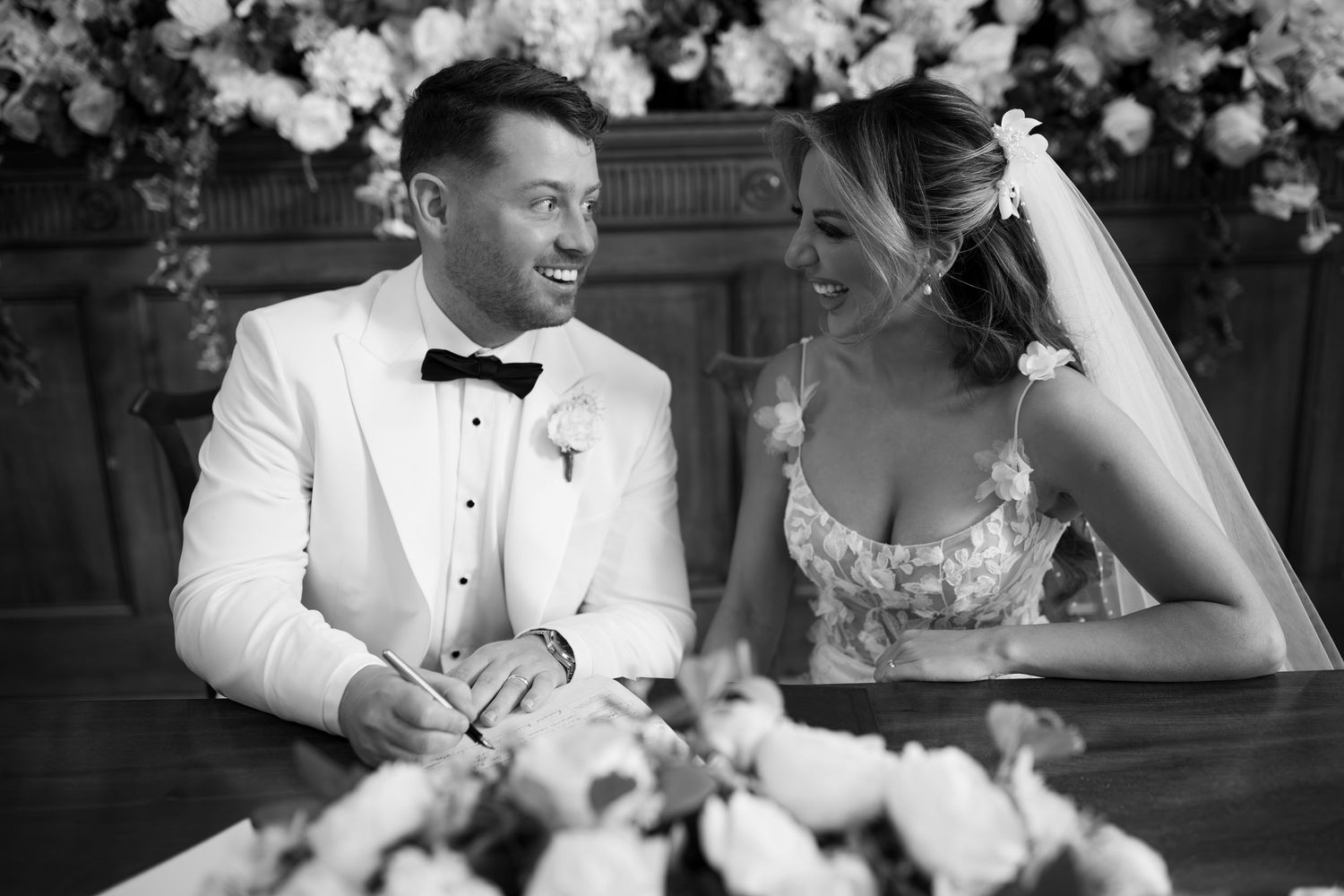 Black and white wedding photo of smiling bride and groom signing marriage register surrounded by floral arrangements.