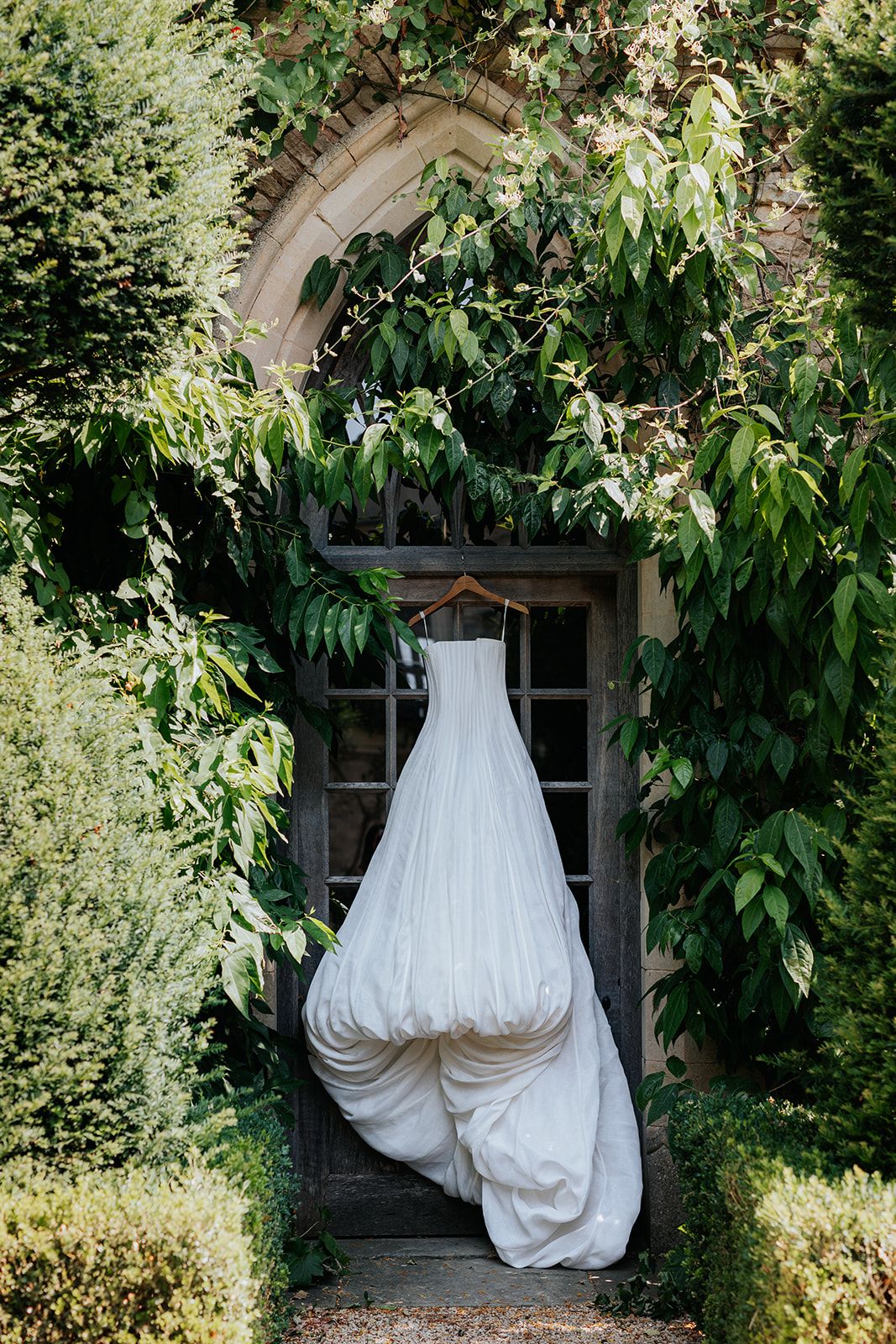 A white wedding dress hanging on a wooden door framed by lush green ivy at an elegant garden venue.