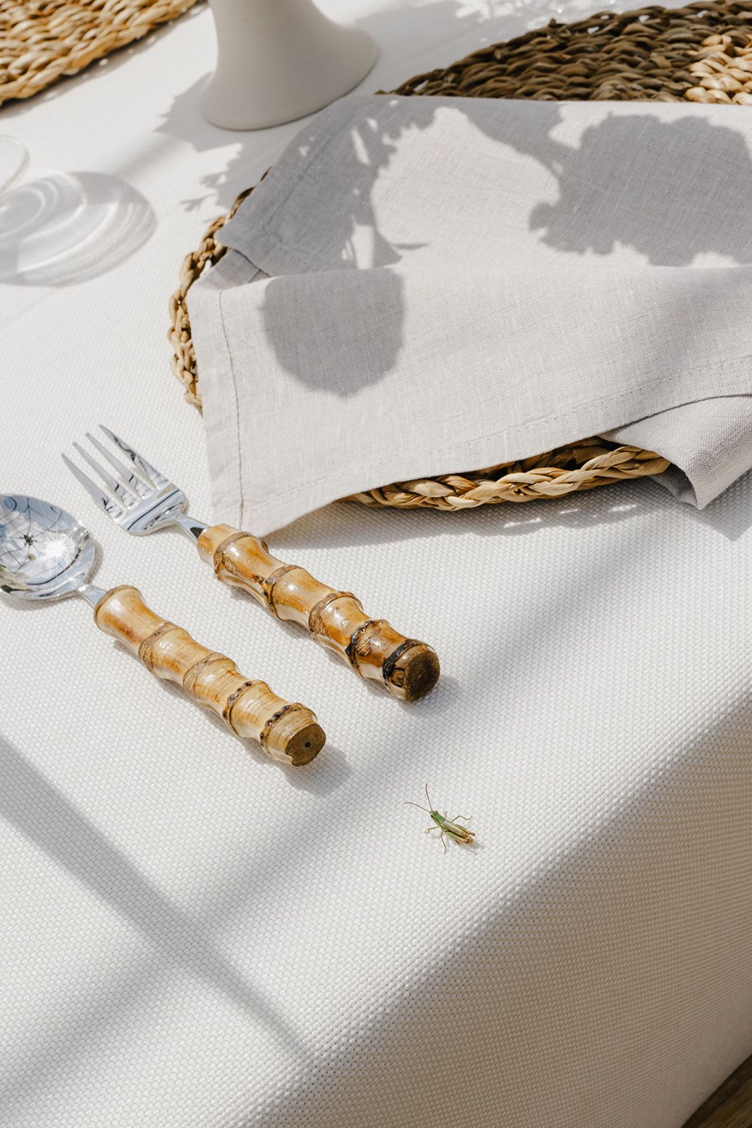 Elegant table setting with bamboo cutlery, linen napkin, wicker placemat, and a grasshopper on white tablecloth.