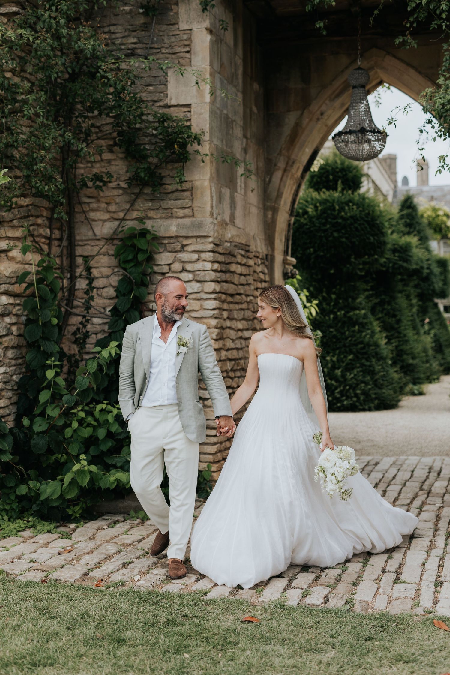 Bride and groom walk hand-in-hand past a stone arch covered in ivy, bride holding white bouquet in elegant strapless gown.