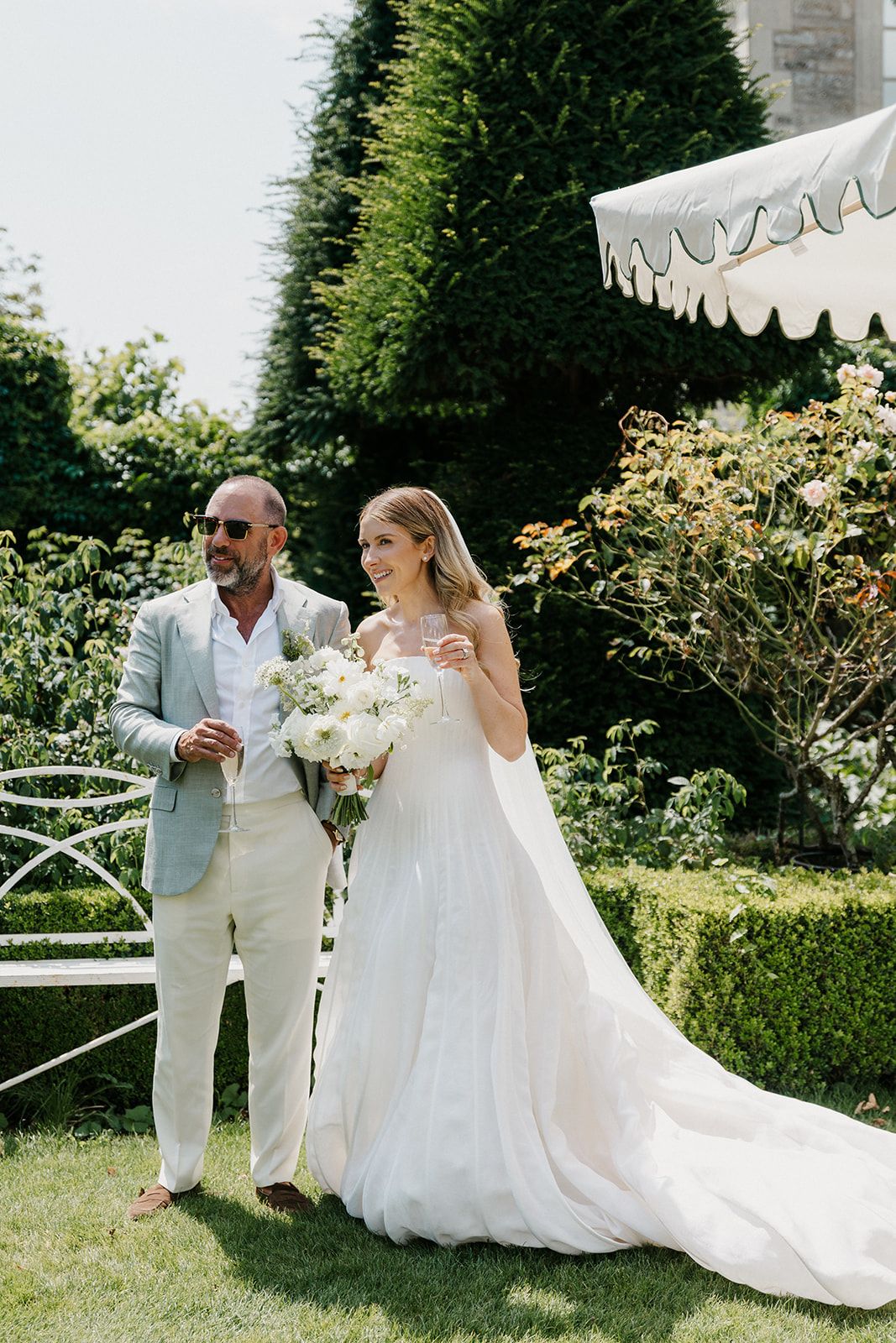 Bride in white gown holding bouquet stands with groom in light suit in lush garden wedding setting.