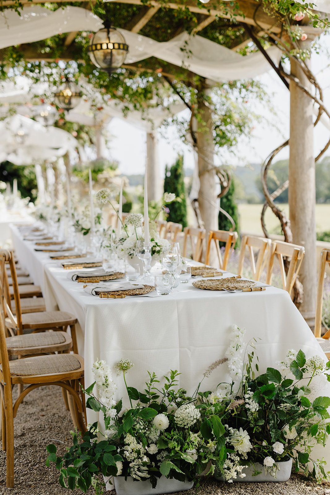 Elegant outdoor wedding reception with long white-draped table, gold chargers, wooden chairs, and lush white floral arrangements.