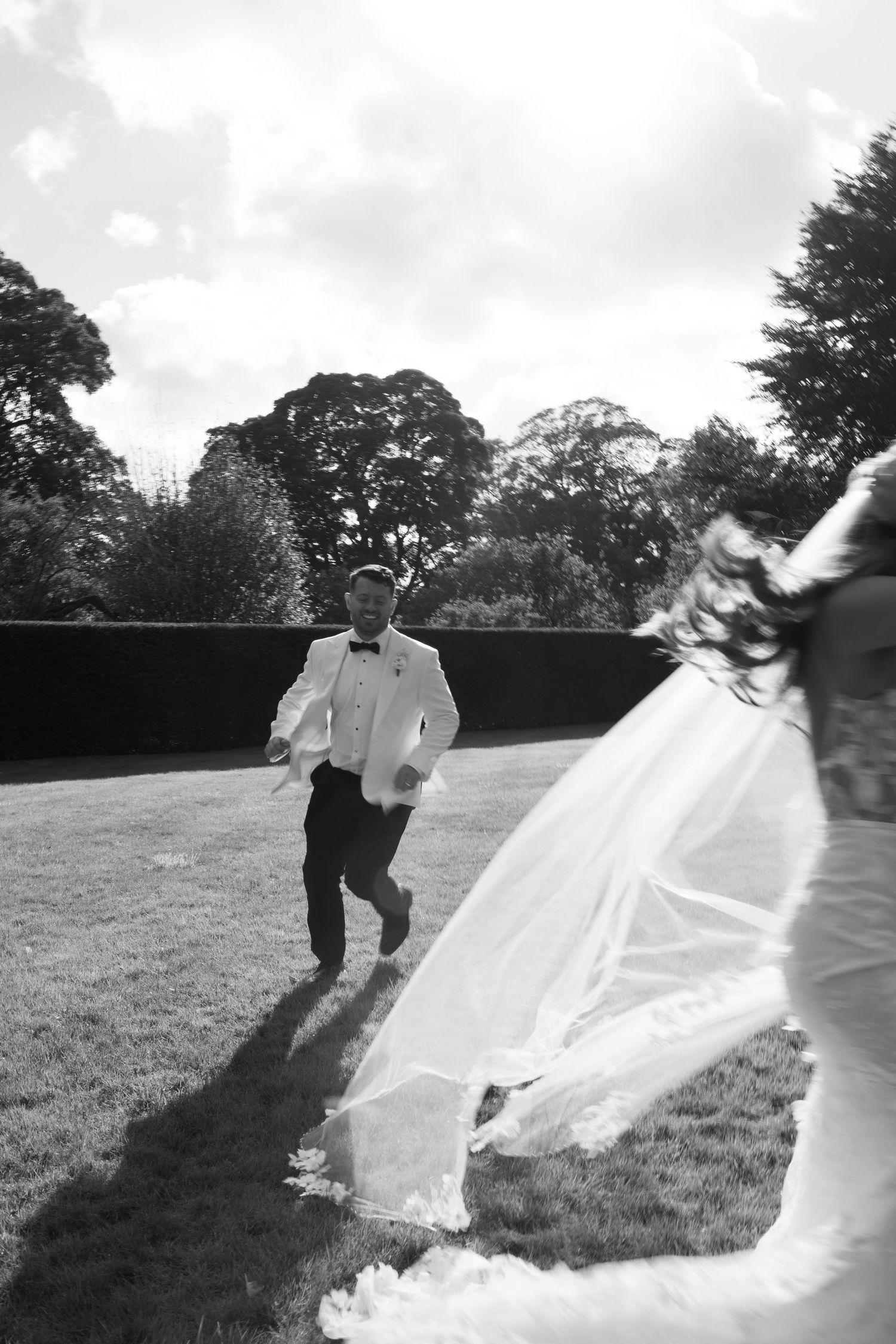 Black and white photo of a groom in white tuxedo running toward bride in flowing wedding dress on lush garden lawn.