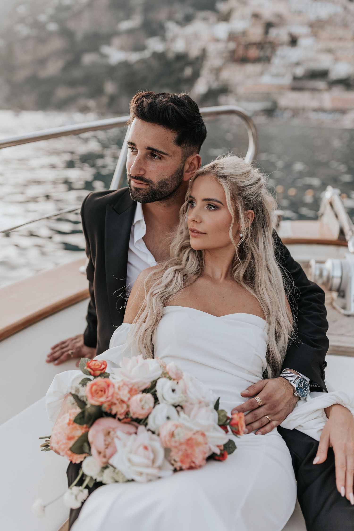 A couple in formal attire stand together on a boat, the woman holding a bouquet of flowers in Positano