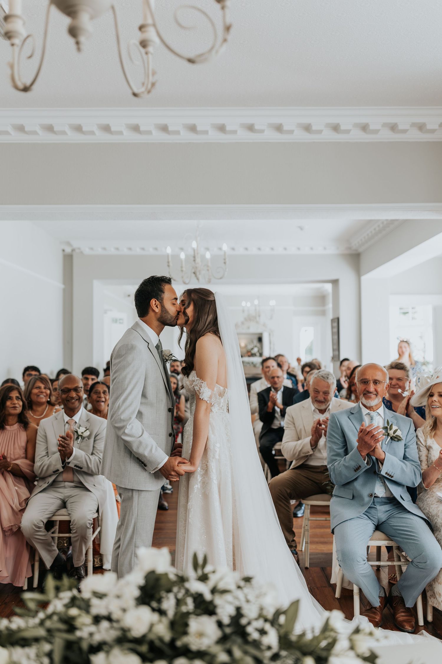 Bride and groom share first kiss at elegant indoor wedding ceremony while guests applaud in bright white venue.