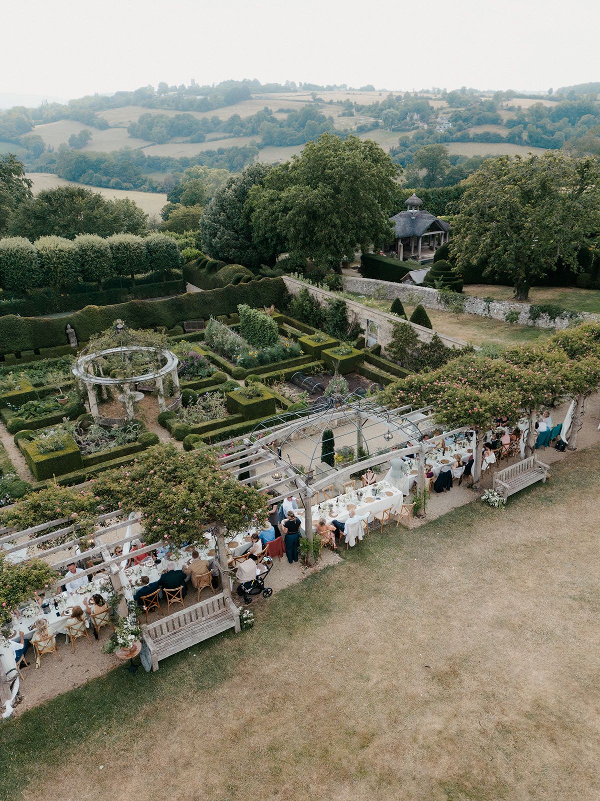 Aerial view of an elegant outdoor wedding reception in a formal English garden with manicured hedges and rolling hills.