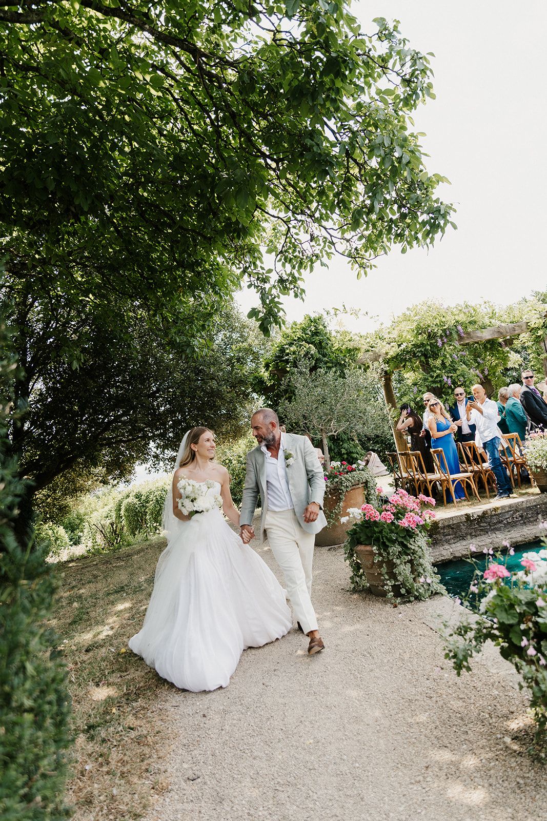 Bride and groom walk hand-in-hand through a sunlit garden path after their outdoor wedding ceremony.