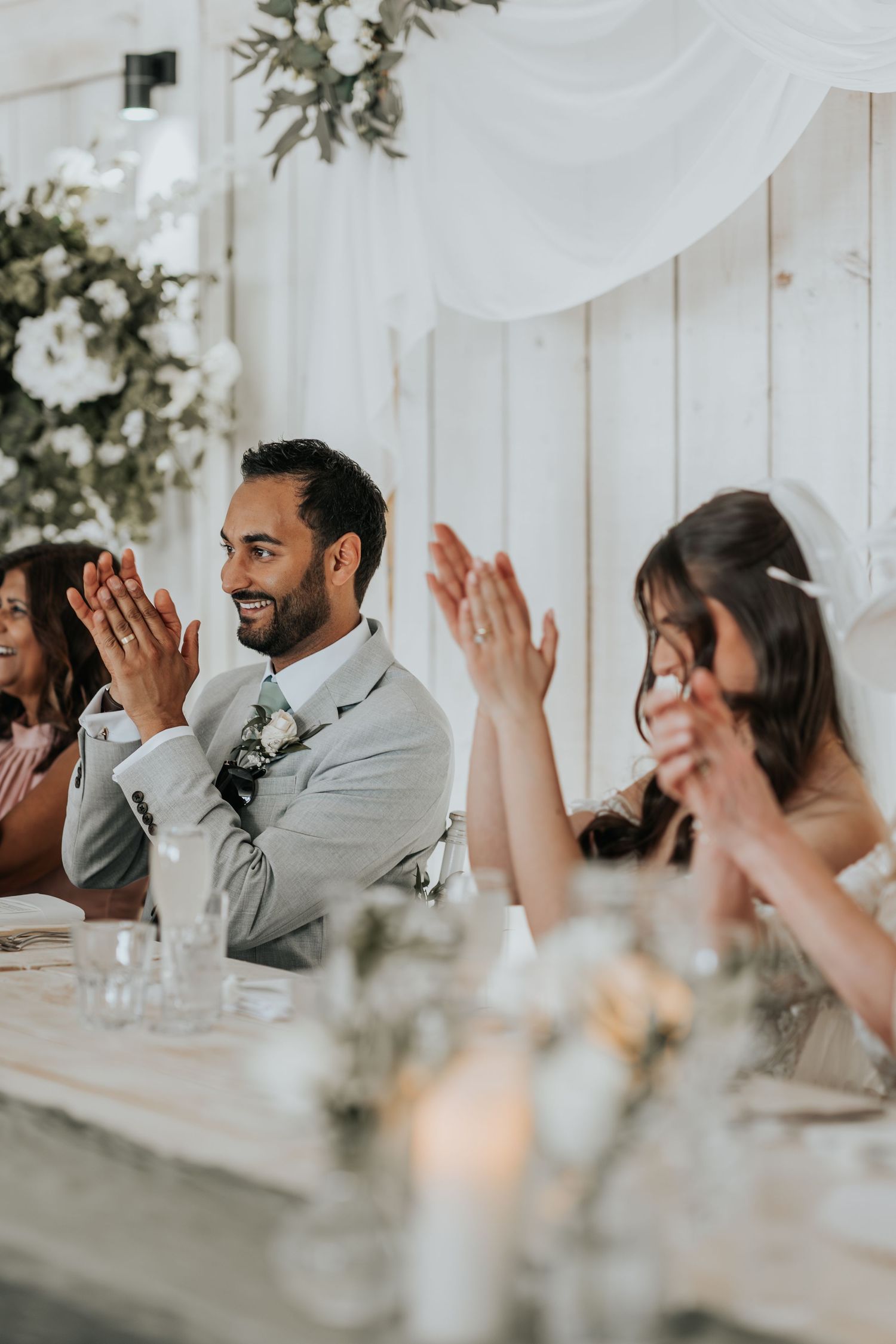 Groom in gray suit clapping at wedding reception table with bride and guests, surrounded by floral decor.