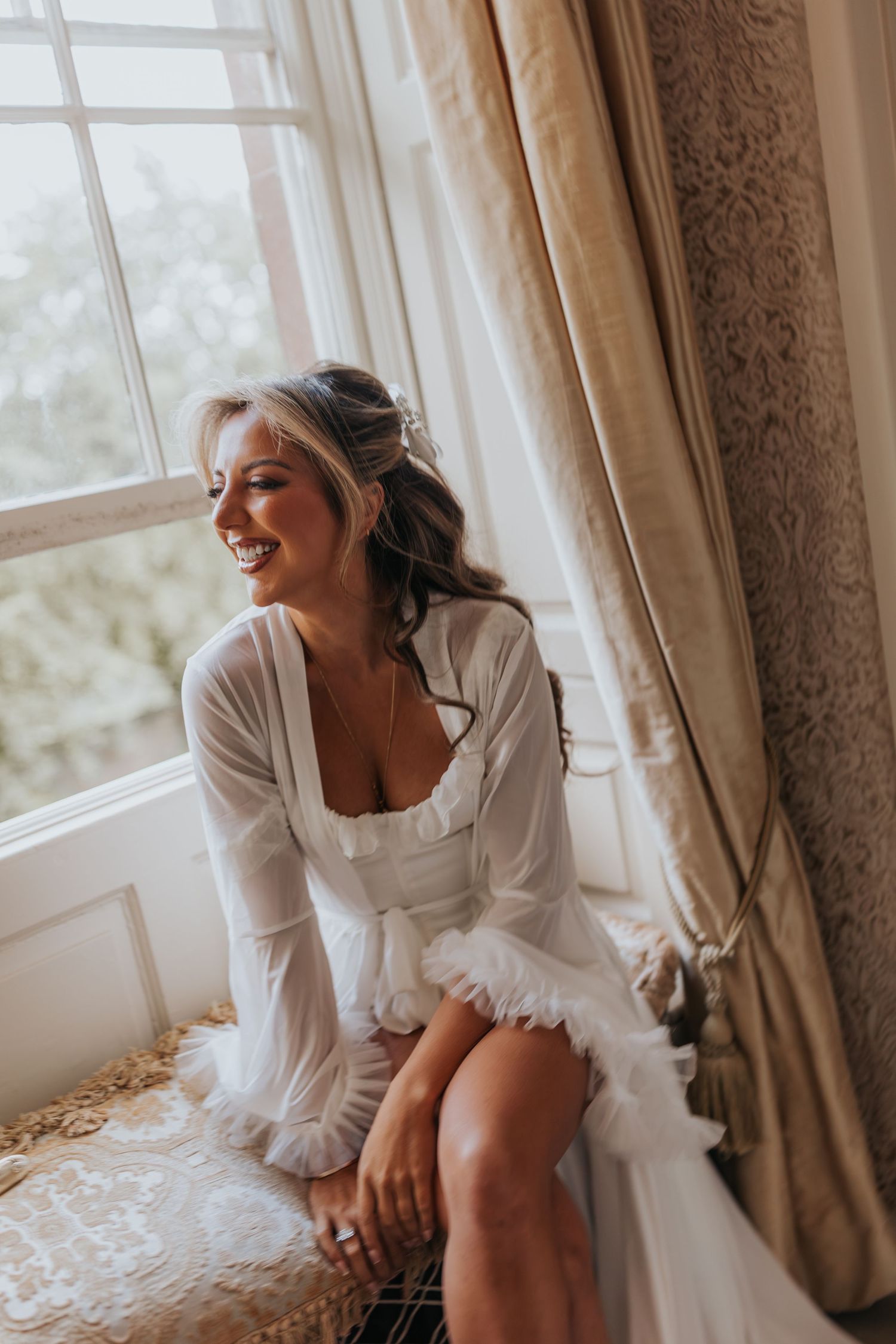 Smiling bride in white robe sitting on window seat with elegant curtains, natural light illuminating her wedding morning preparations.