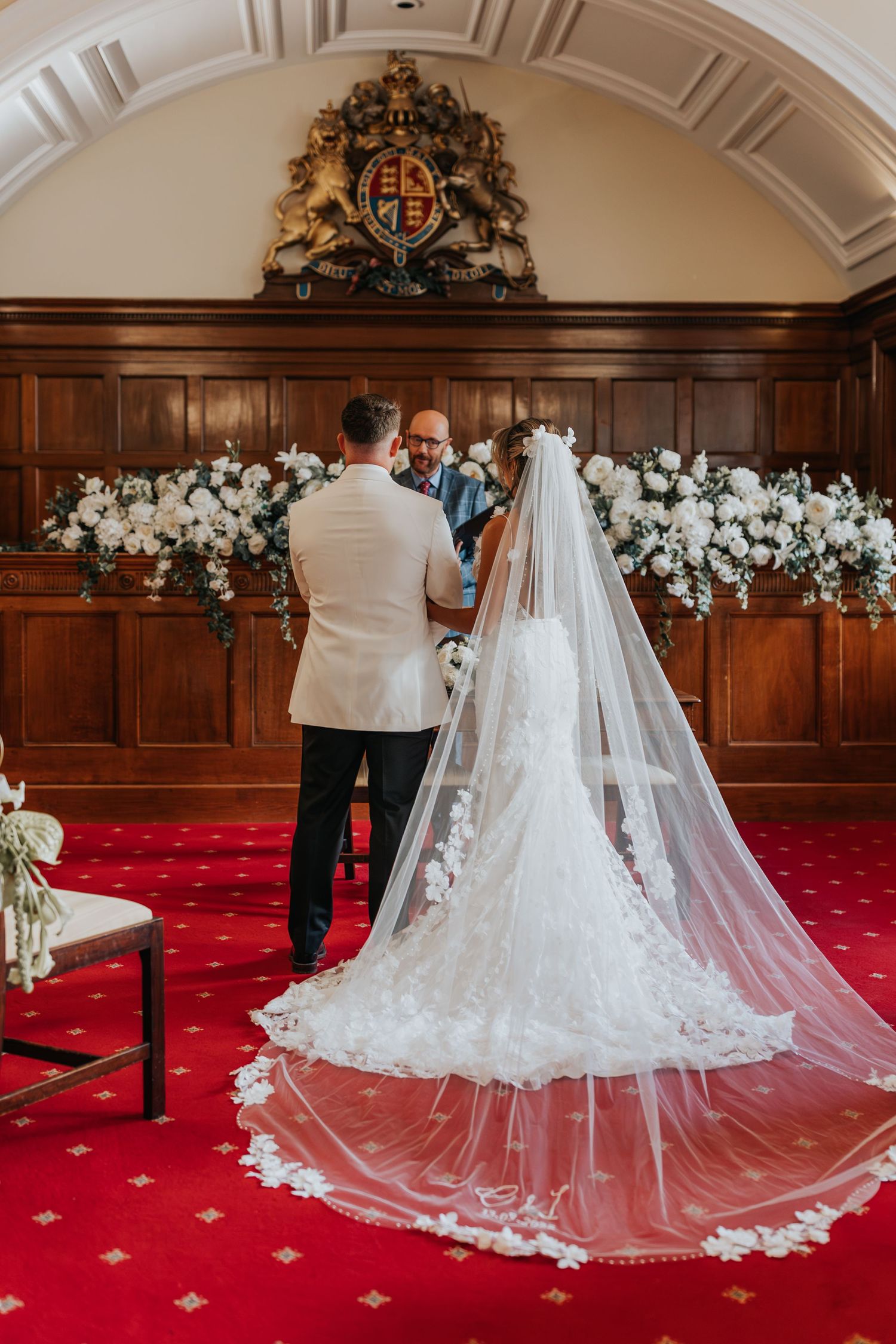 Bride and groom stand at the altar during a wedding ceremony in an elegant wood-paneled room with white floral arrangements.