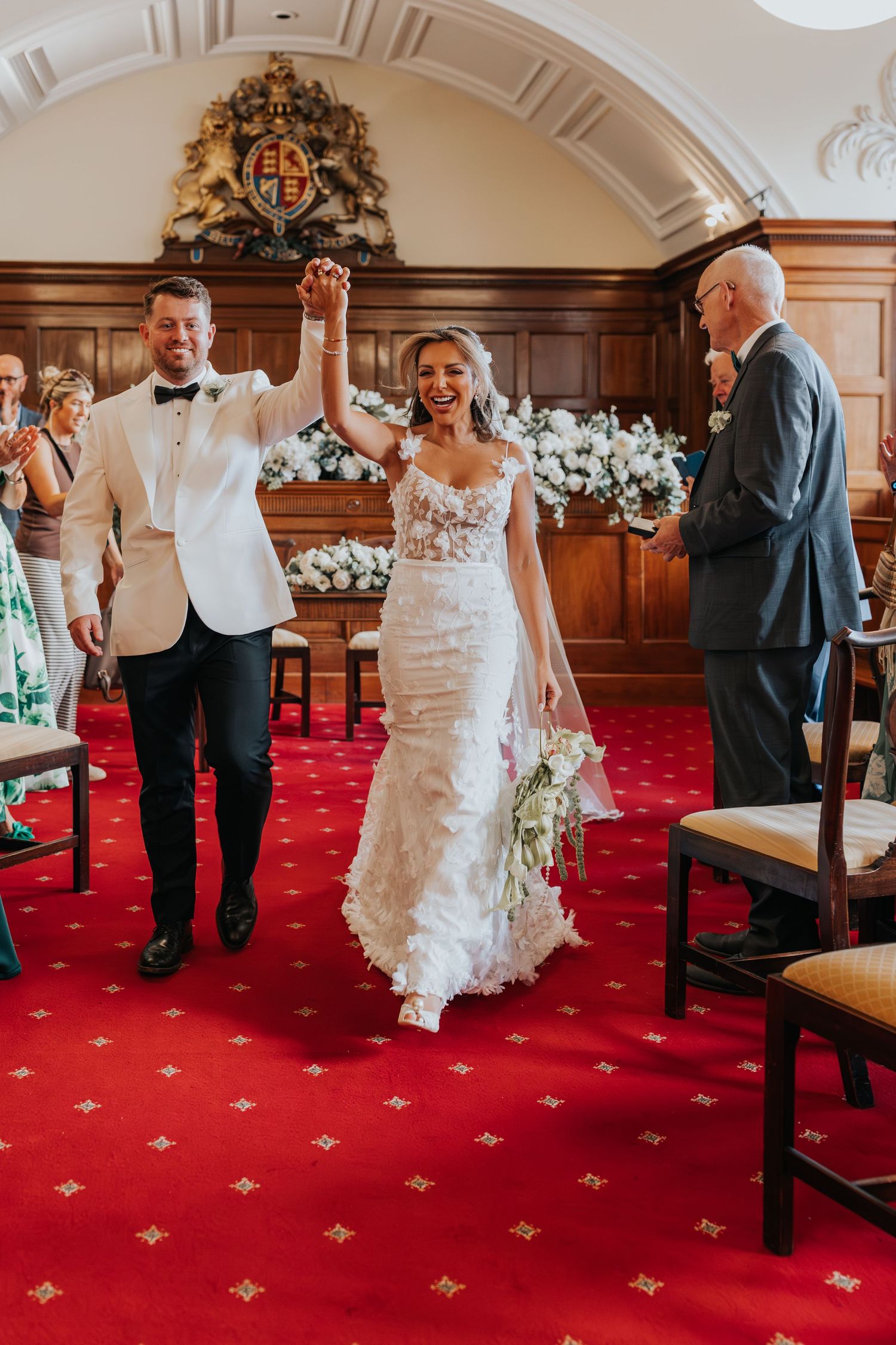Joyful newlyweds raising hands walking down red carpet aisle in elegant wedding ceremony venue.
