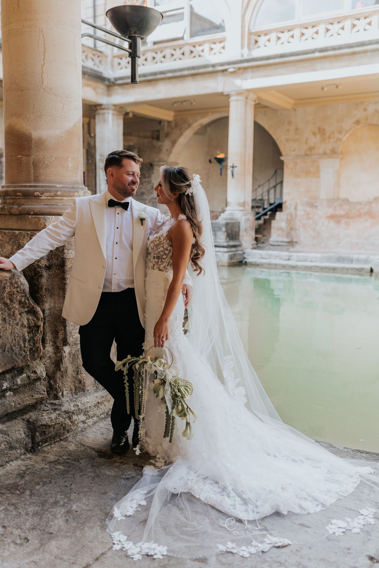 Bride and groom pose romantically beside the Roman Baths pool, she in lace gown with veil, he in white tuxedo jacket.