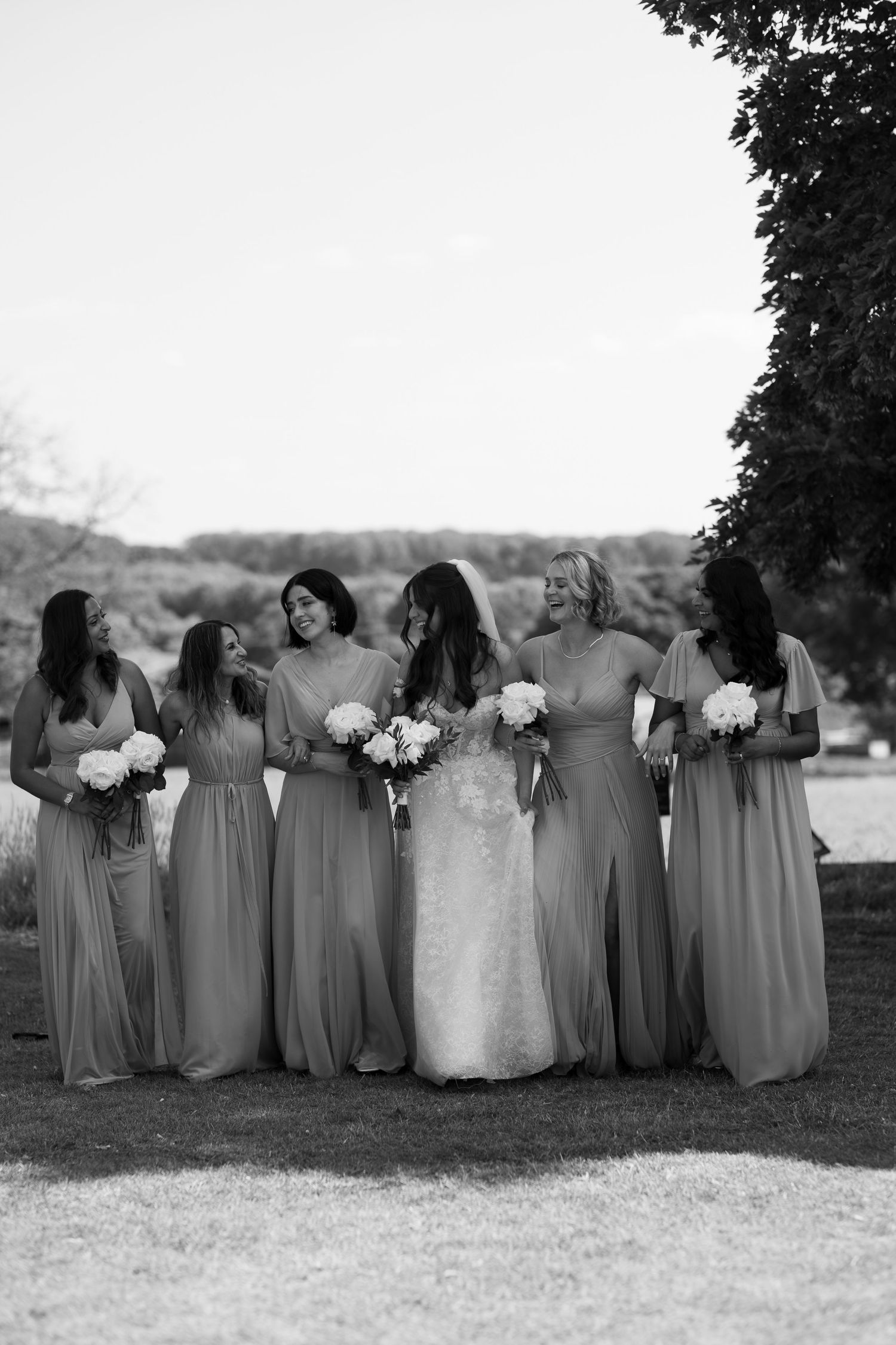 A group of women in long gray dresses stand together holding bouquets of flowers in a grassy outdoor setting.