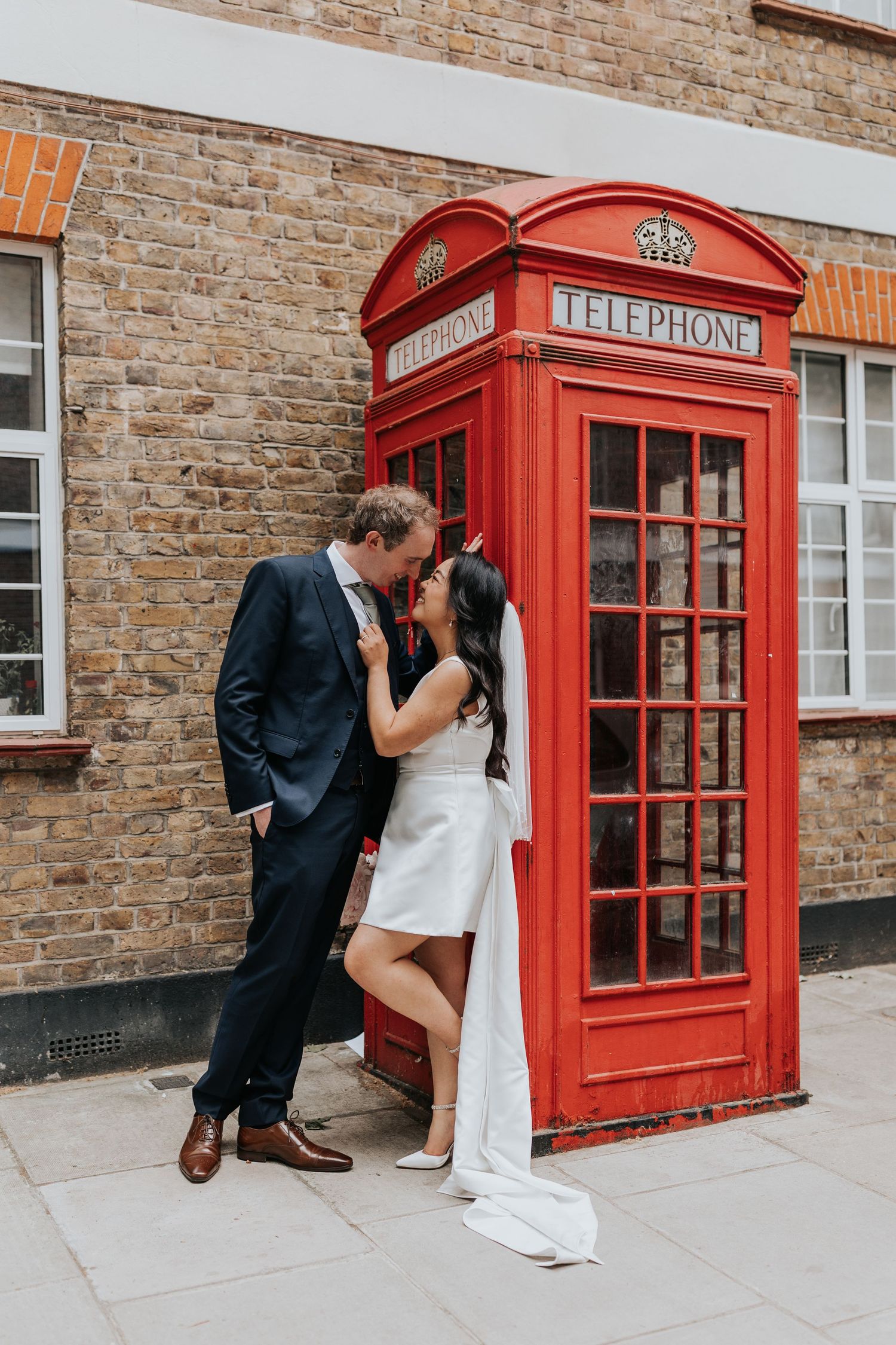 A bride and groom embrace in front of a classic red telephone booth, creating a romantic and charming scene.