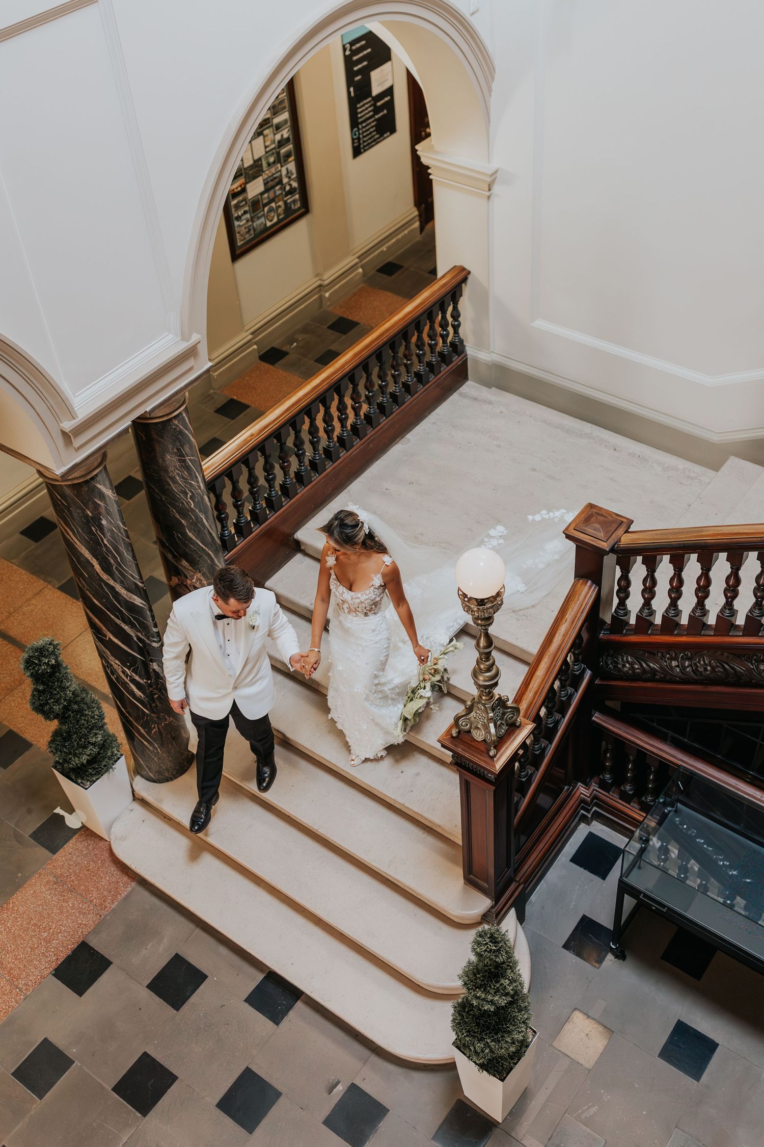 Bride and groom walking down elegant grand staircase in historic venue, aerial view of wedding couple holding hands.