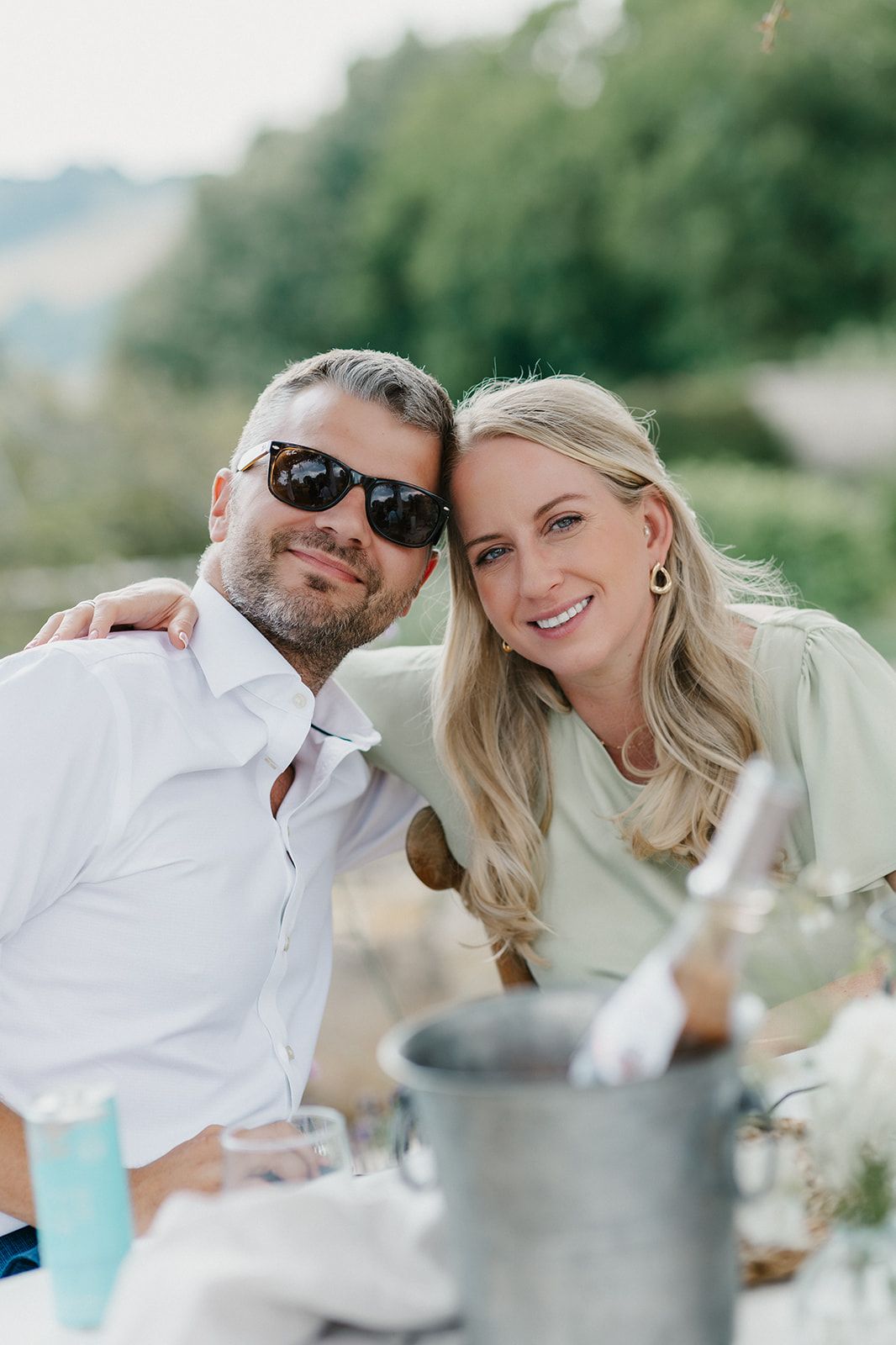 A smiling couple poses outdoors at a picnic table with a champagne bucket, man in sunglasses and white shirt.
