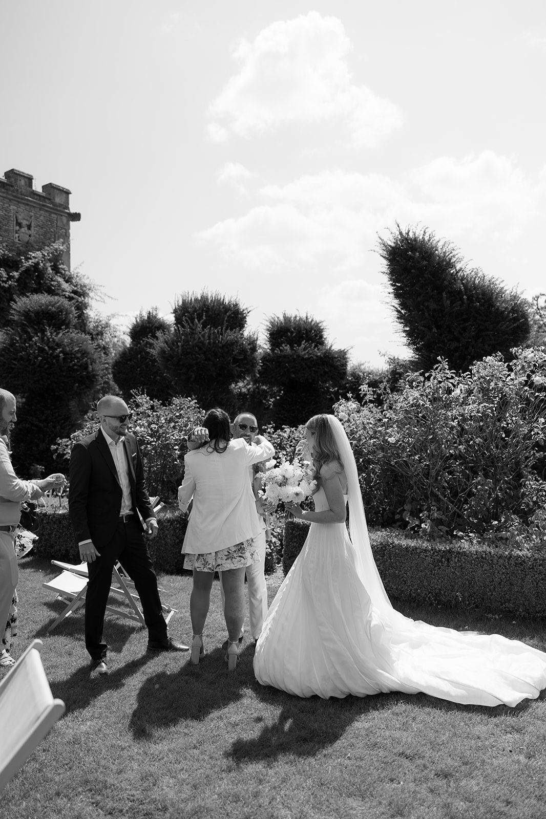 Black and white photo of bride in garden with wedding guests and castle backdrop.
