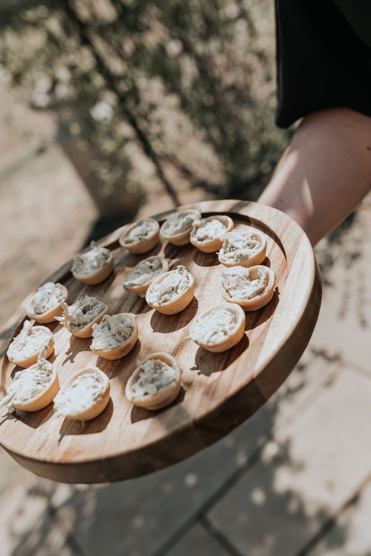 Person holding a wooden oval board with cream cheese crostini appetizers served outdoors at an event.