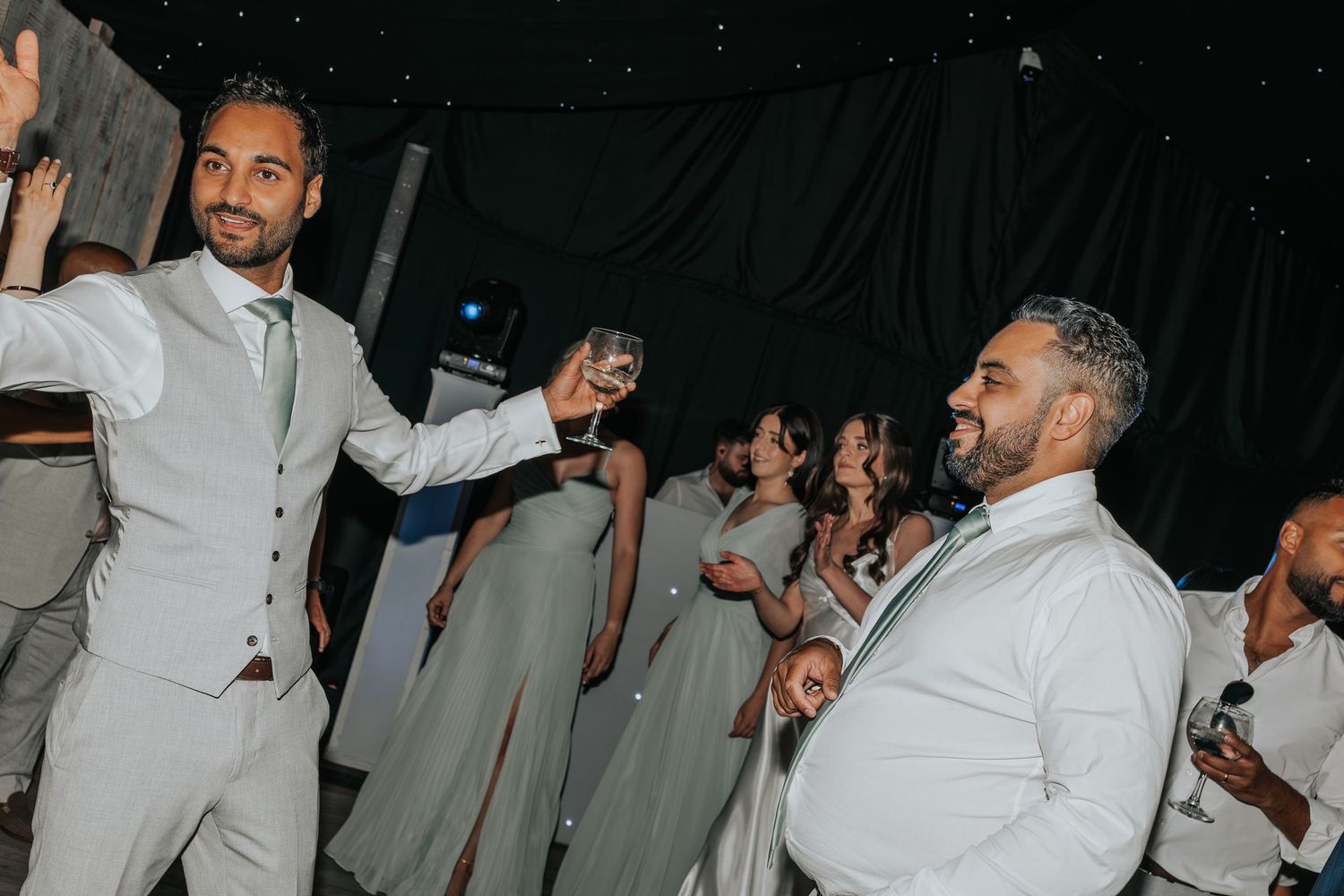 Men in white suits toasting with champagne glasses at a wedding reception dance floor with bridesmaids in sage green.