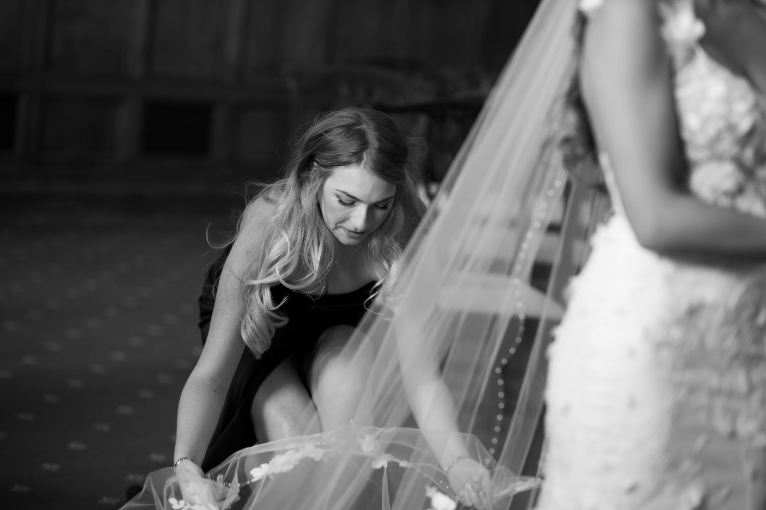 Bridesmaid in black dress adjusting bride's wedding veil in elegant black and white photo.