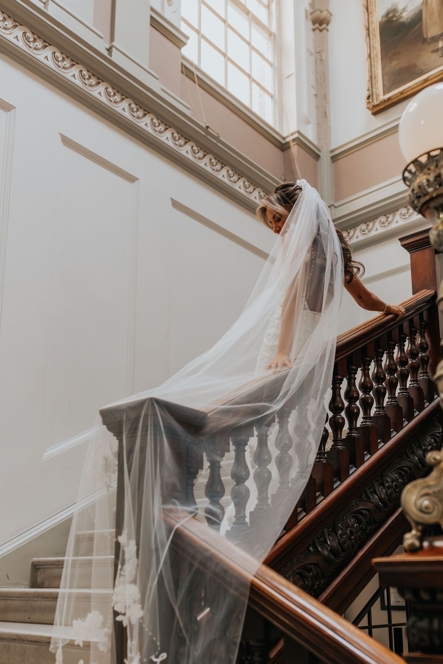 Bride in a white veil descends an elegant wooden staircase in a grand venue with ornate moldings.